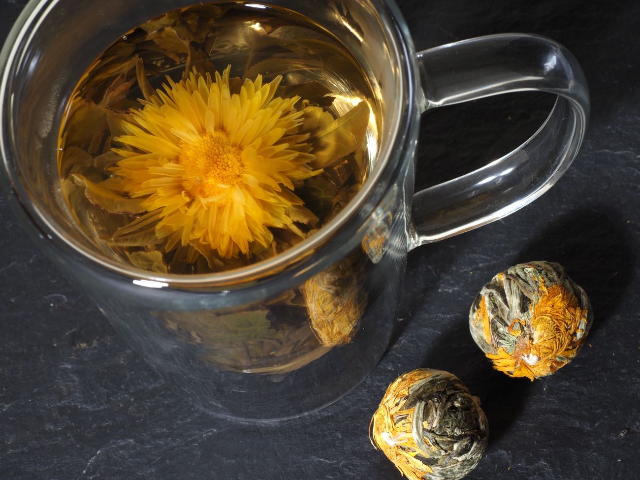 Glasbecher mit aufgeblühter Teeblume. Zwei Teeblumen daneben. Auf schwarzem Untergrund.
