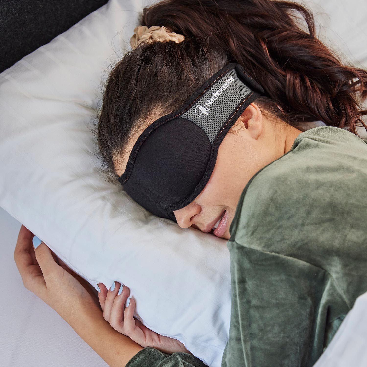 Frau mit schwarzer Schlafmaske im Bett. Maske bedeckt Augen, graues Band mit Logo.
