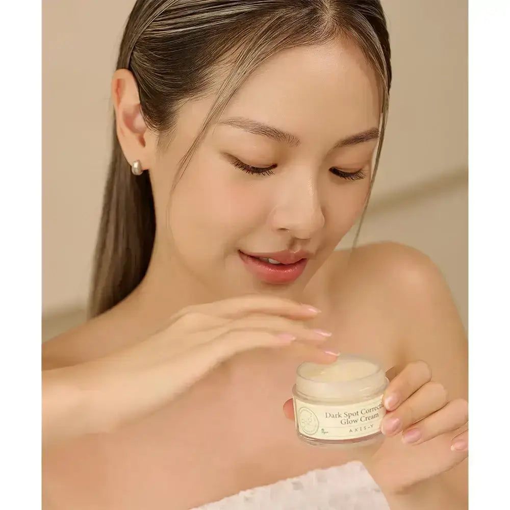 Frau hält Tiegel AXIS-Y Dark Spot Correcting Glow Cream. Helle Haut, lange Haare. Produkt im Vordergrund.