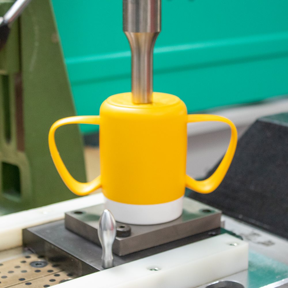 Gelber Becher mit zwei Henkeln, weißer Rand. Wird von einer Maschine bearbeitet.