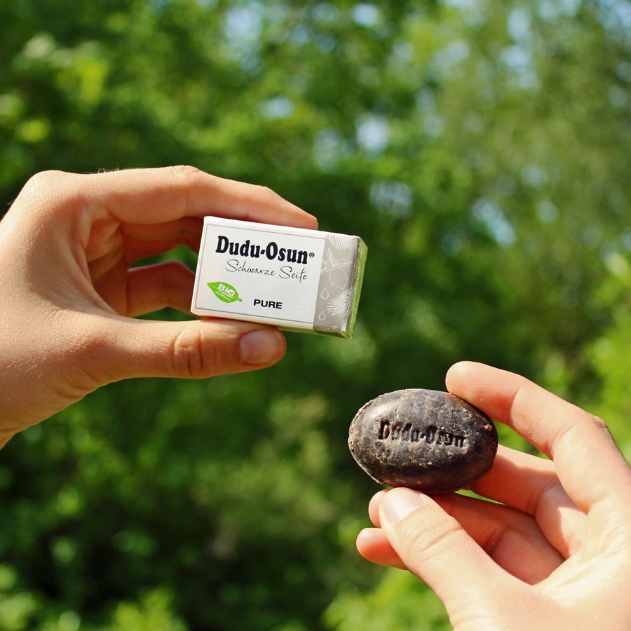 Hände halten Dudu-Osun-Seife und Verpackung. Schwarze Seife, ovale Form, mit Logo. Verpackung mit Schriftzug.