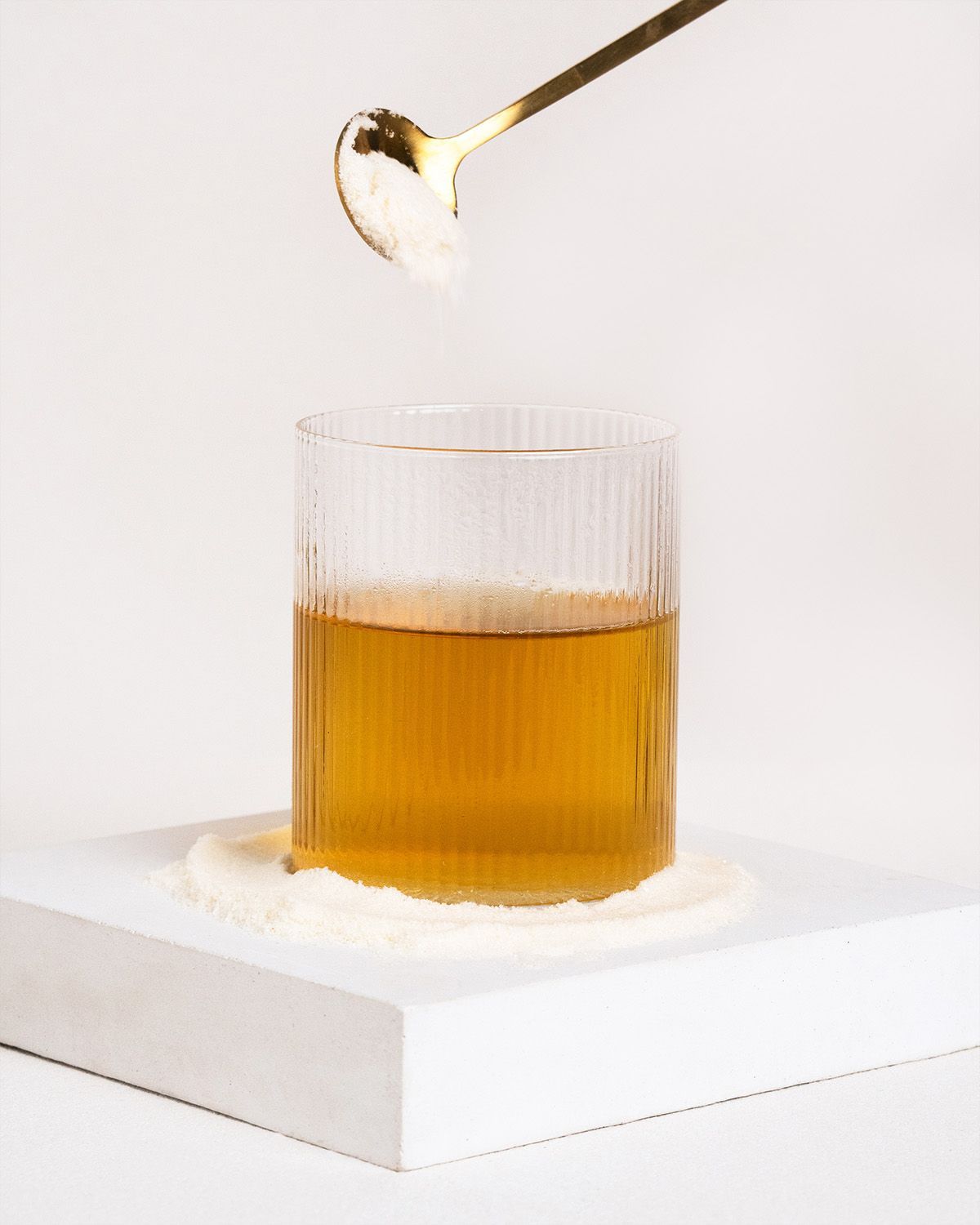Glas mit Flüssigkeit, Pulver auf weißem Podest, goldener Löffel mit Pulver darüber.