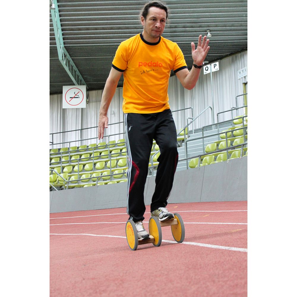 Mann auf gelbem Koordinationstrainer. Gelbe Räder, Holzkonstruktion. Logo auf T-Shirt. Sportanlage im Hintergrund.