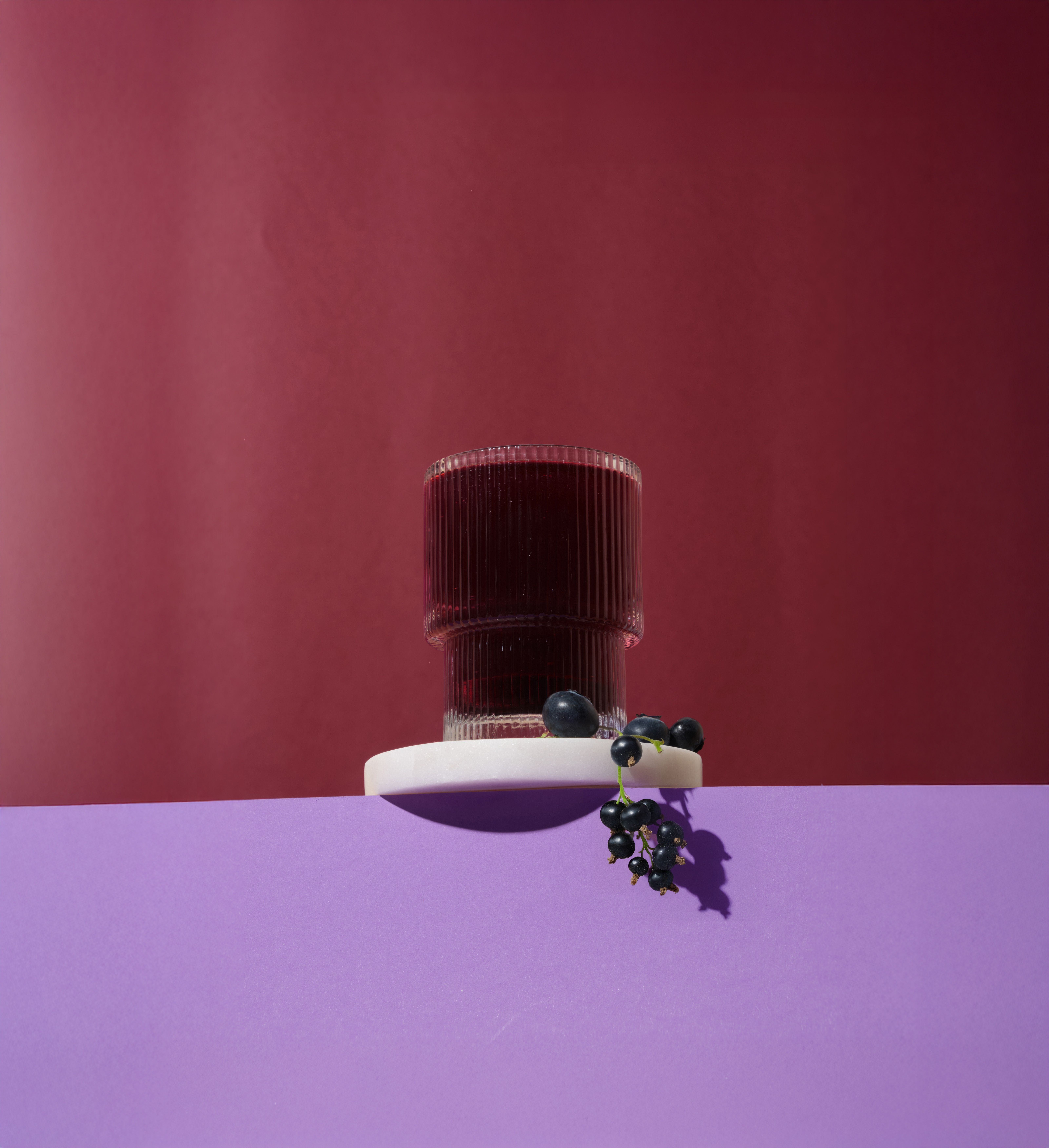 Glas mit dunkler Flüssigkeit auf weißem Podest. Blaubeeren und Johannisbeeren daneben. Hintergrund: Lila und Rot.