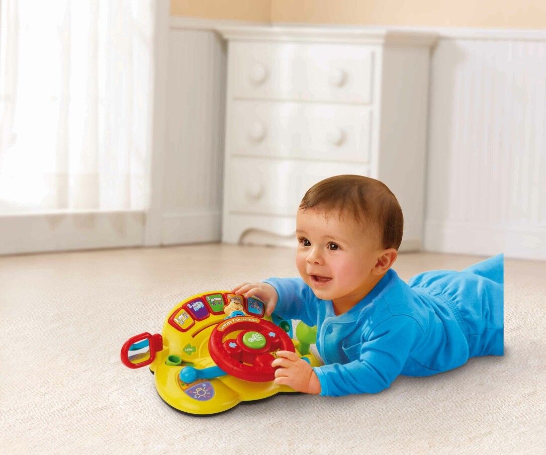 Baby liegt auf dem Bauch und spielt mit gelbem Spielzeug-Lenkrad. Rotes Lenkrad, gelbe Basis.