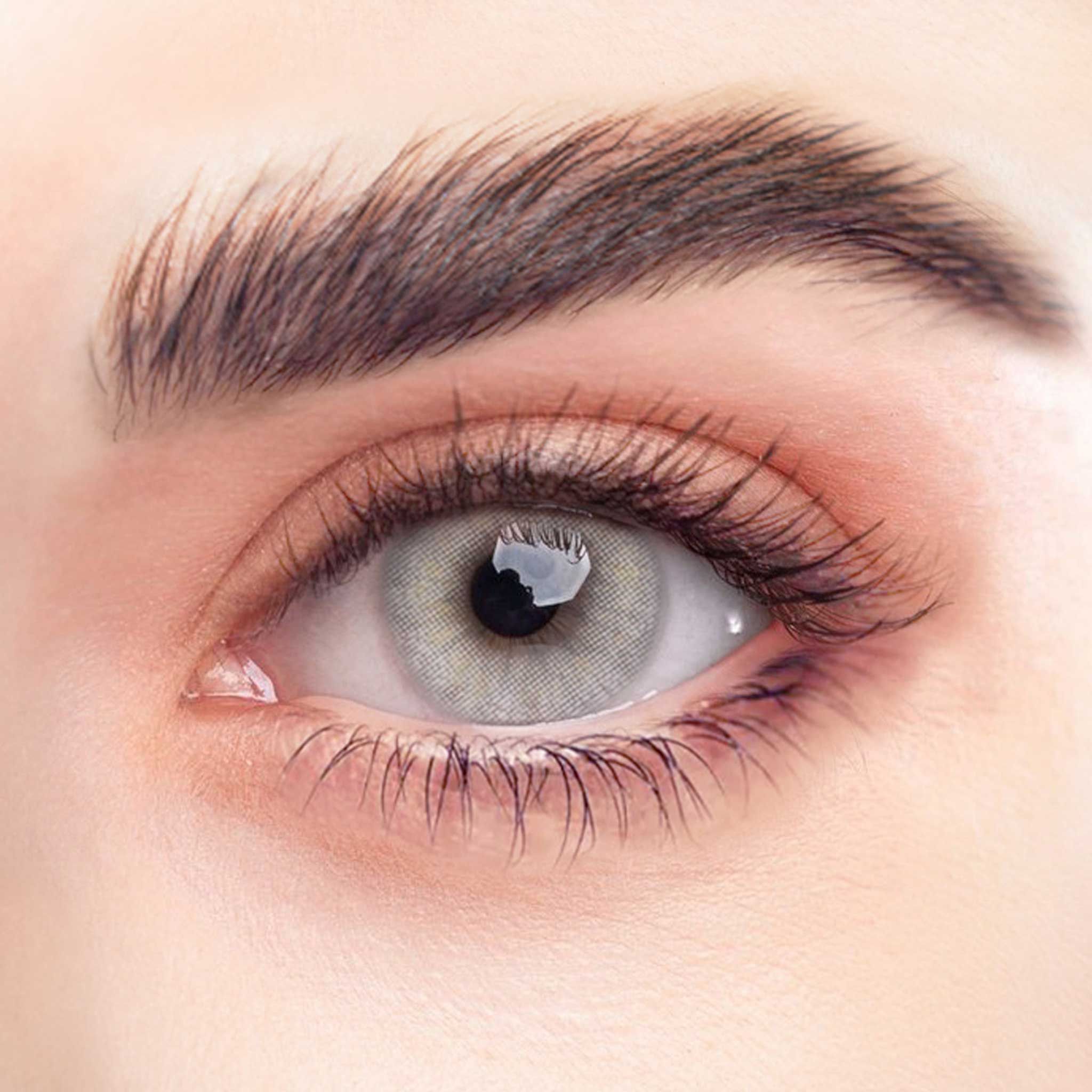 Nahaufnahme eines Auges mit grauer Kontaktlinse. Sichtbar: Iris, Pupille, Wimpern, Augenbraue. Die Linse hat einen hellgrauen Farbton und reflektiert das Licht.