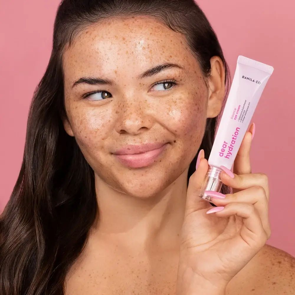 Frau hält rosa Tube. Aufschrift: BANILA CO, Dear Hydration, Bounce Eye Cream. Hintergrund: rosa. Frau lächelt.
