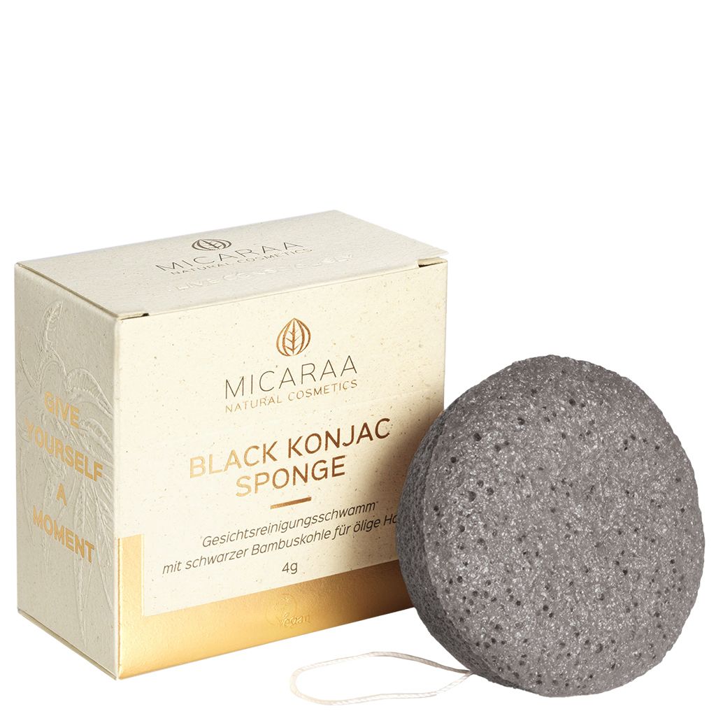 Grauer, runder Schwamm neben Verpackung. Verpackung mit Logo und Text: BLACK KONJAC SPONGE.