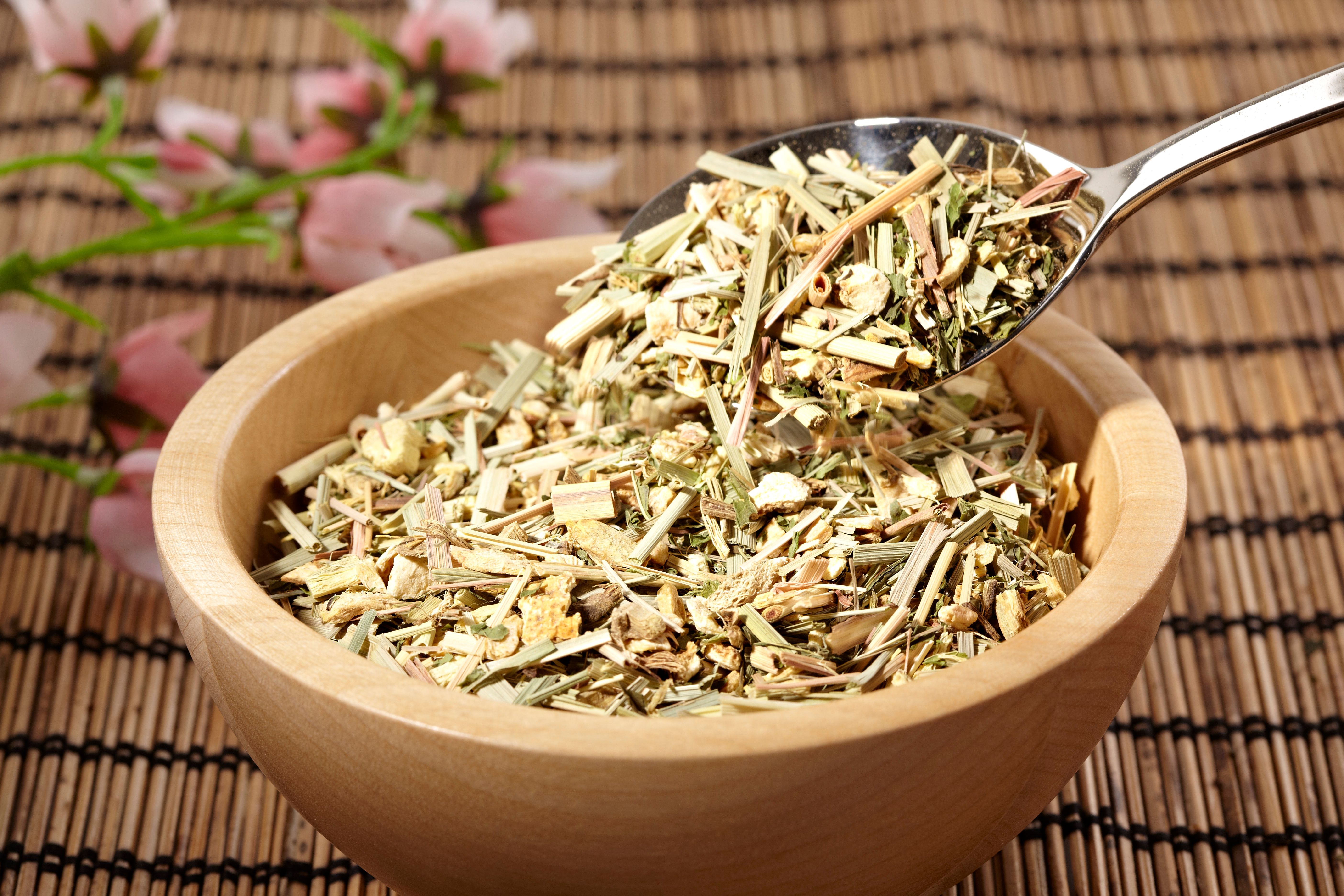 Holzschale mit Kräutermischung. Ein Löffel hebt Kräuter aus der Schale. Hintergrund: Bambusmatte und Blüten.