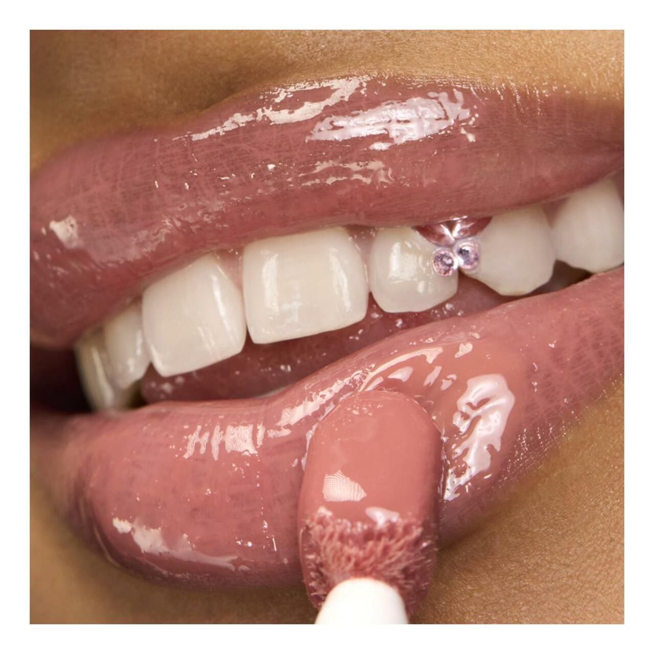 Nahaufnahme von Lippen mit aufgetragenem Lipgloss. Der Applikator ist sichtbar. Zähne mit Schmuck.
