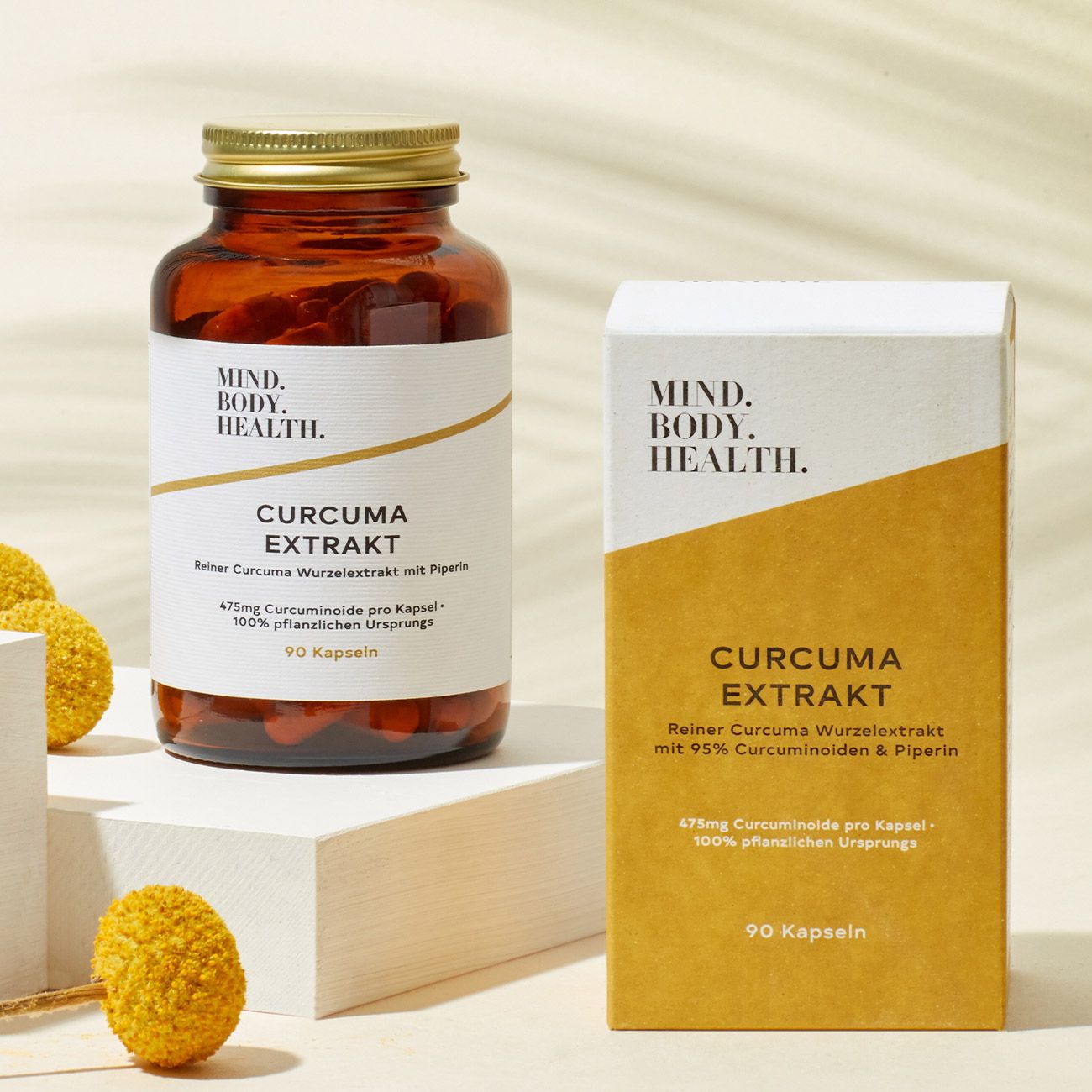 Braune Glasflasche und Schachtel mit "MIND.BODY. HEALTH.", "CURCUMA EXTRAKT". Beide mit Inhaltsangaben. Auf weißem Podest.