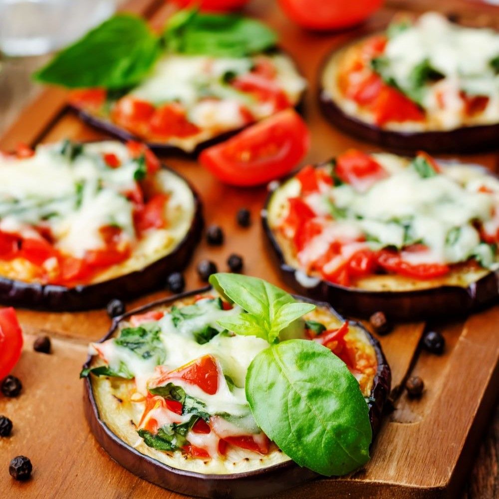 Auberginenscheiben mit Tomaten, Käse und Basilikum auf Holzbrett. Tomaten und Pfefferkörner daneben.