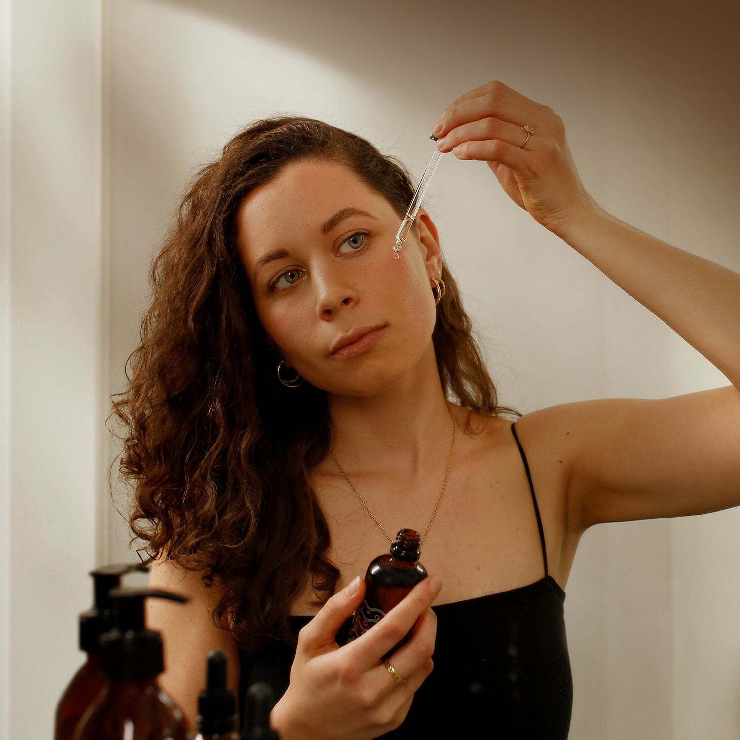 Frau hält eine braune Flasche und einen Tropfer. Sie tropft Öl auf ihre Haut. Im Hintergrund Spiegel und weitere Flaschen.