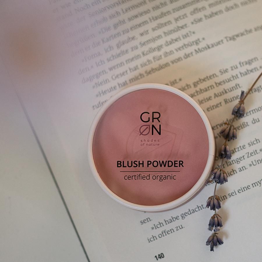 Runder, rosa GRÜN Blush Powder Behälter auf Buch. Text: BLUSH POWDER, certified organic. Weißer Rand.