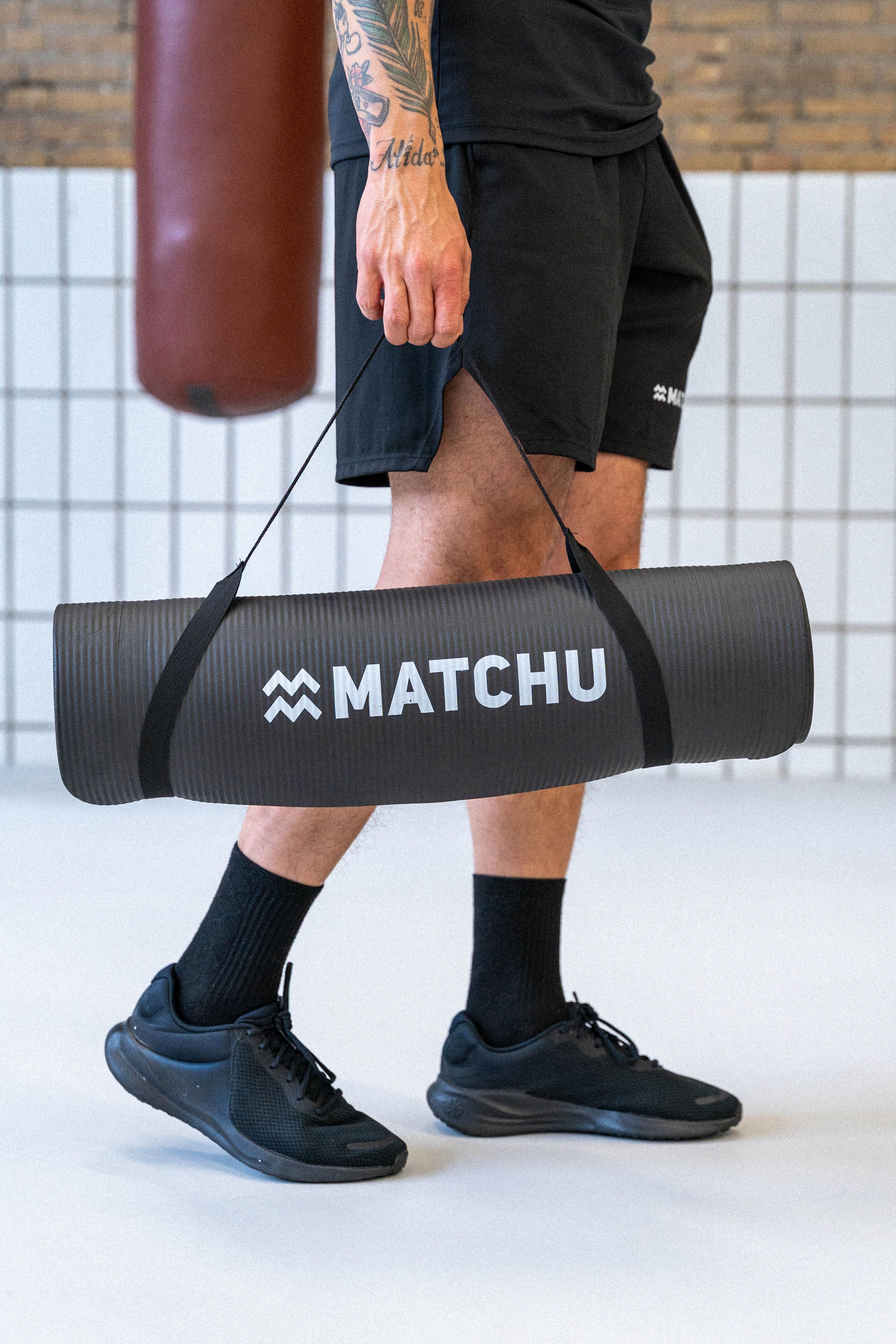 Person trägt gerollte schwarze Yogamatte mit "MATCHU"-Logo. Schwarze Kleidung.