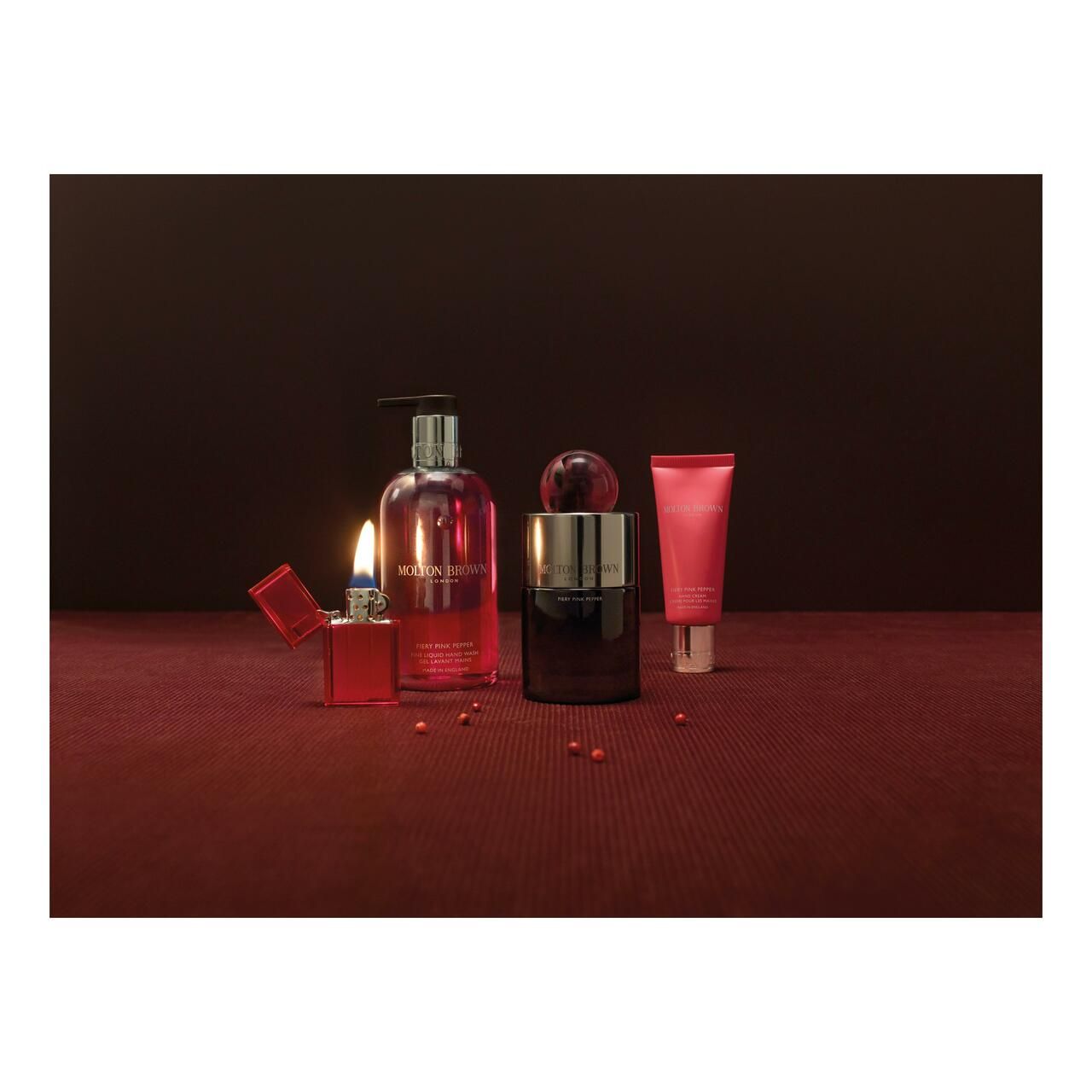 Produkt-Arrangement: Flakon, Handseife, Creme, Feuerzeug. Auf rotem Untergrund. Molton Brown, Fiery Pink Pepper.