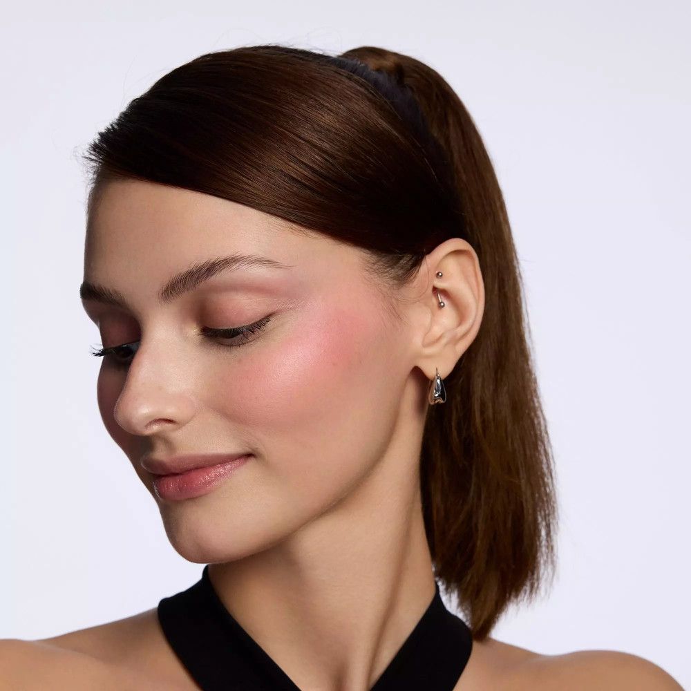 Porträt einer Frau mit rosa Rouge auf den Wangen. Sie trägt ein schwarzes Oberteil und Ohrringe. Ihr Haar ist zurückgebunden.