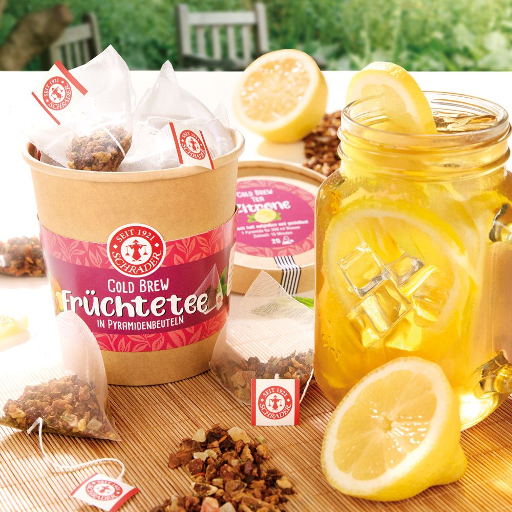 Becher mit Teebeuteln, Glas mit Getränk und Zitrone. Aufschrift: Schrader Cold Brew Früchtetee Zitrone. Teebeutel mit Logo.