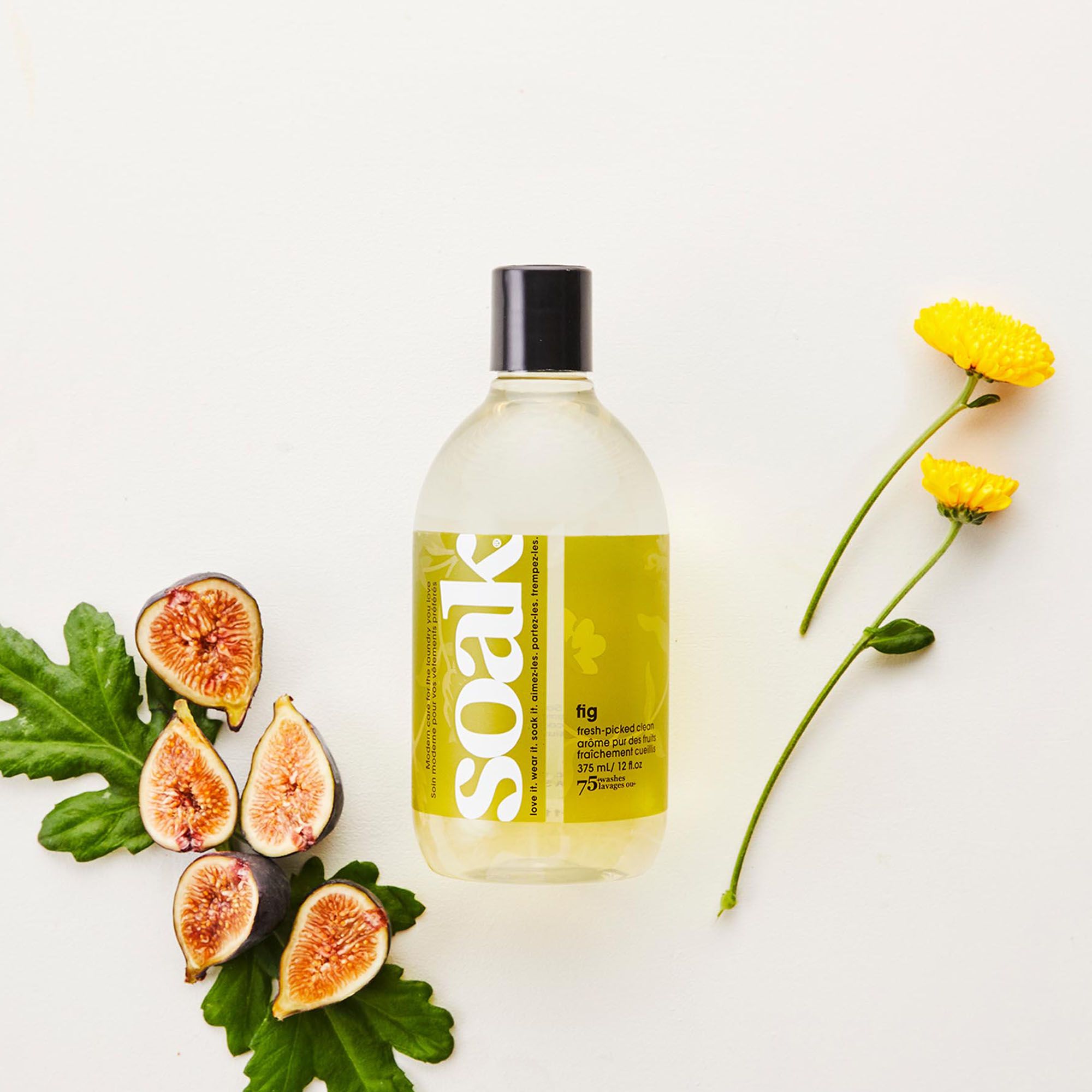 Klare Flasche mit schwarzem Deckel, gelbem Etikett mit "Soak" und "fig". Daneben Feigen und gelbe Blüten.
