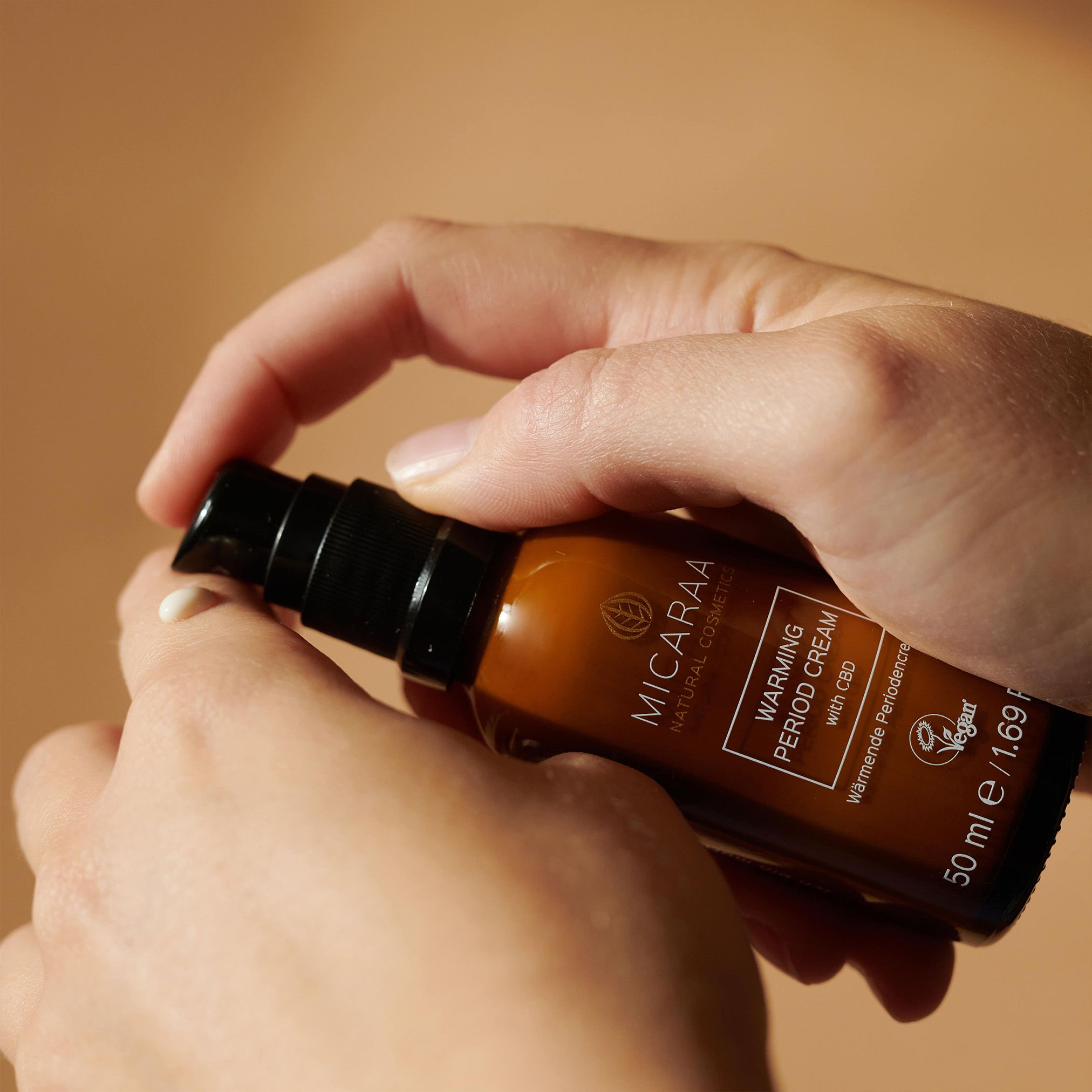 Hände drücken Creme aus brauner Flasche. Aufschrift: MICARAA Warming Period Cream with CBD. 50 ml.