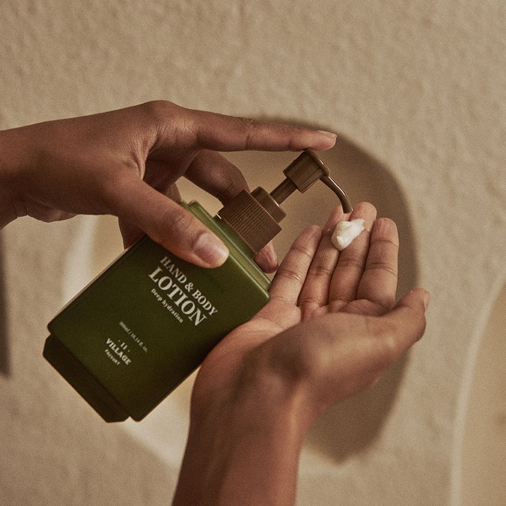 Hände drücken Lotion aus grüner Flasche. Flasche mit braunem Pumpkopf. Text: Hand & Body Lotion, 11 Village Factory.