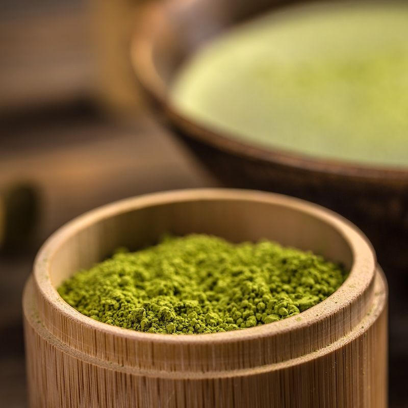 Nahaufnahme von grünem Matcha-Pulver in einem kleinen Holzbehälter, im Hintergrund eine Schale mit Matcha-Tee.
