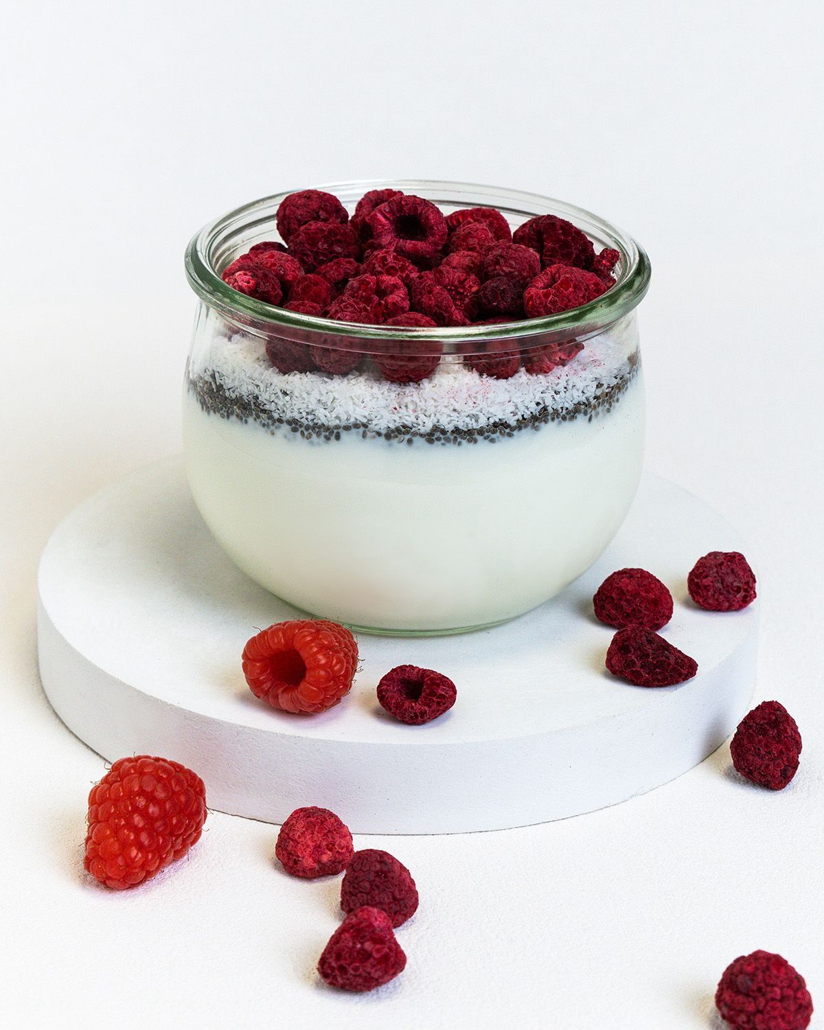 Glas mit Joghurt, Chia-Samen und gefriergetrockneten Himbeeren. Einige Himbeeren liegen auf einer weißen Oberfläche.