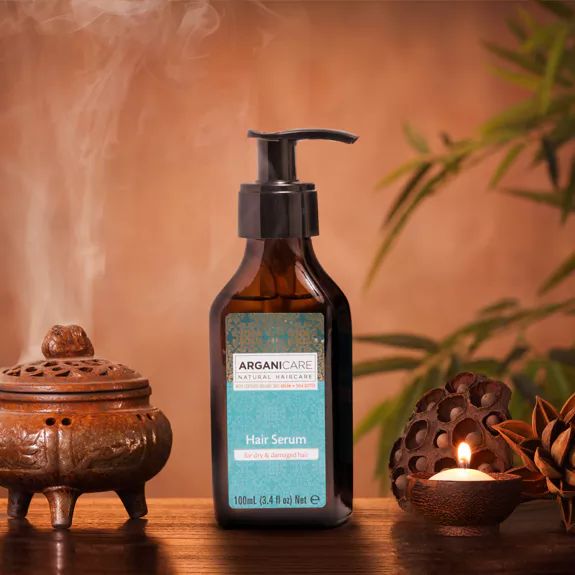 Haarserum-Flasche mit Pumpe. Aufschrift: Arganicare, Hair Serum. Hintergrund: Kerze, Räuchergefäß, Pflanzen.
