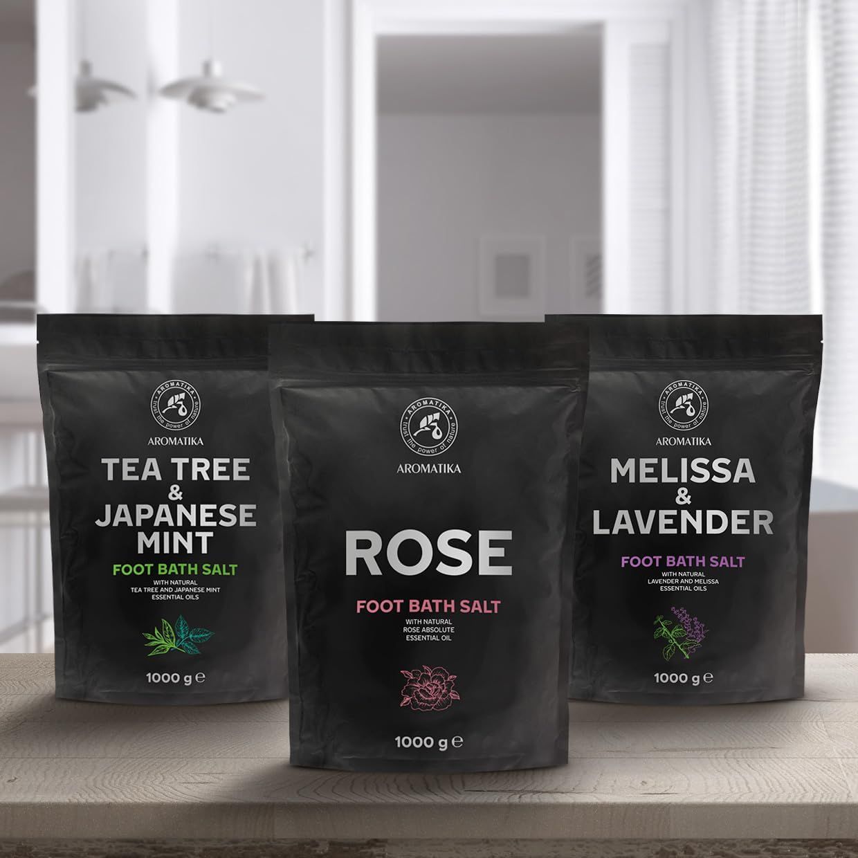 Drei schwarze Beutel Fußbadesalz auf einem Holztisch. Aufschrift: Tea Tree & Japanische Minze, Rose, Melisse & Lavendel. Marke: AROMATIKA. 1000 g.