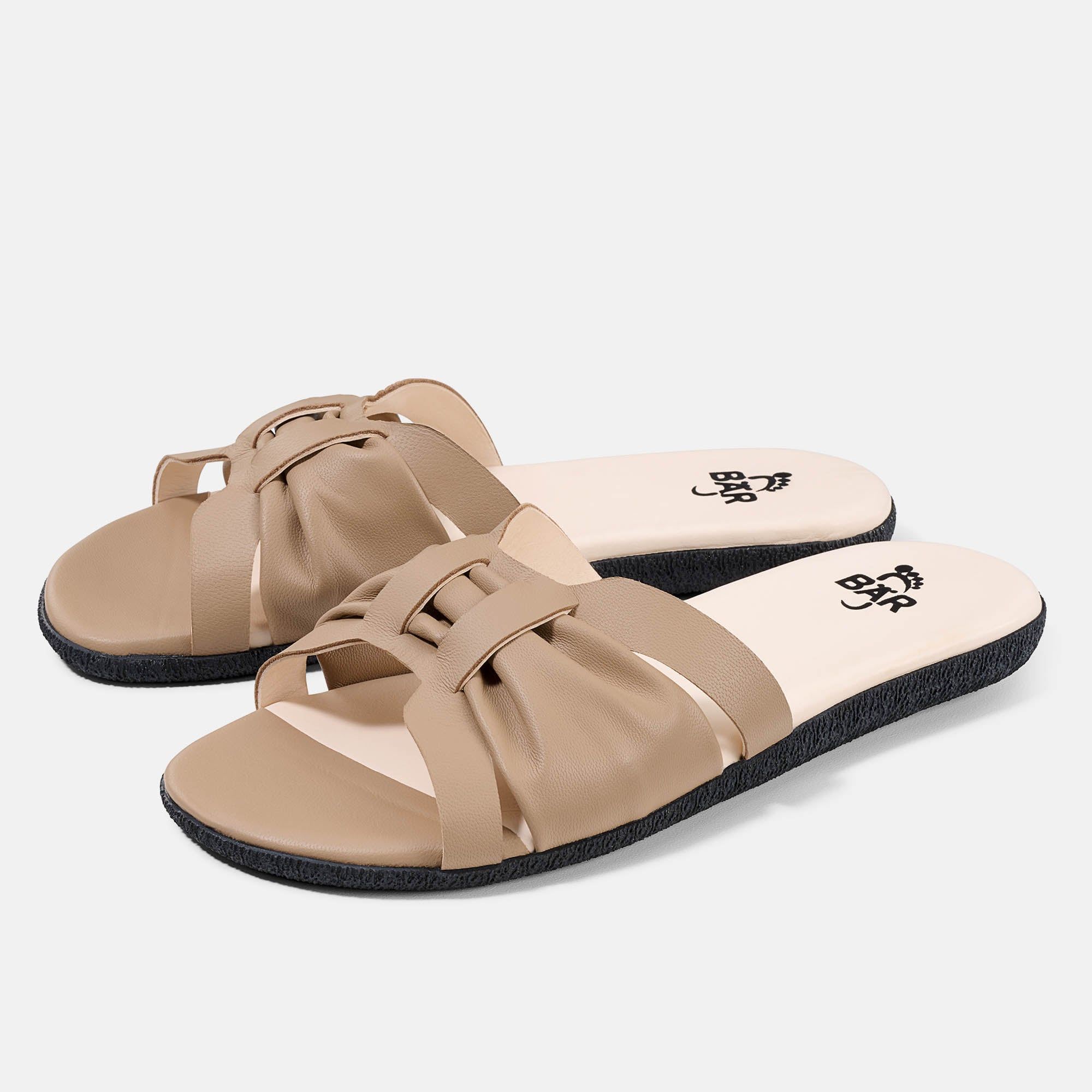 Beige Pantoletten mit breitem Riemen. Schwarze Sohle, helle Innensohle mit Logo. Zwei Schuhe nebeneinander.