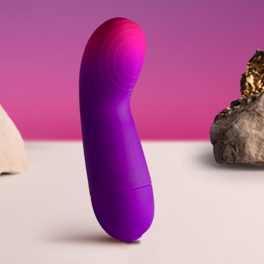 Lila-pinkfarbener Vibrator mit geschwungener Form. Auf weißem Untergrund, mit Steinen im Hintergrund.