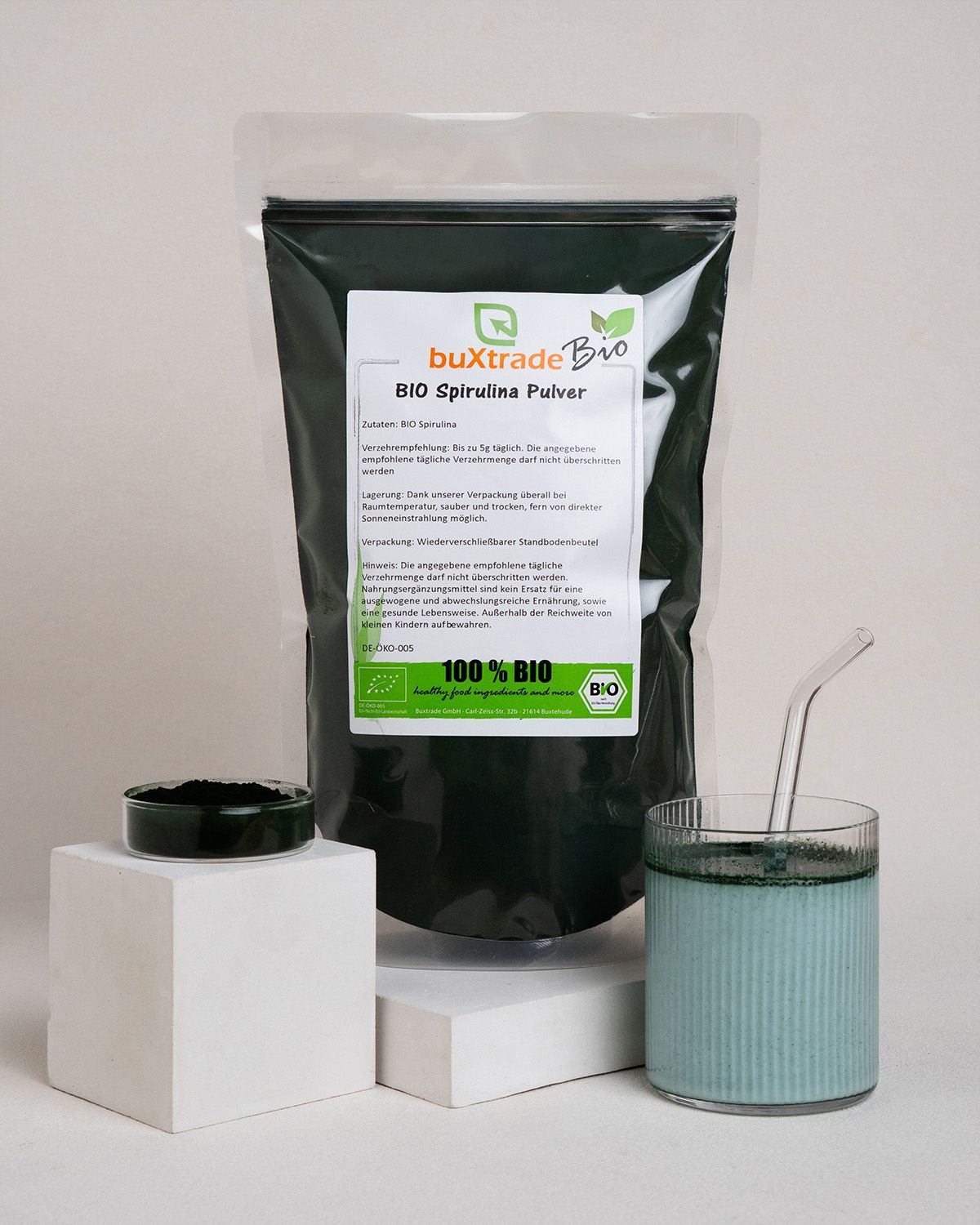 Verpackung mit buXtrade Bio Spirulina Pulver, Glas mit Getränk, Schälchen mit Pulver. Auf weißen Podesten.
