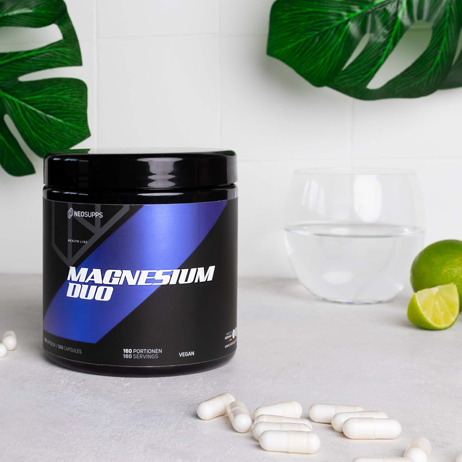 Magnesium Duo Dose. Schwarzer Behälter mit blauem Etikett. Text: Magnesium Duo, Vegan. Kapseln und Glas Wasser auf weißem Untergrund.