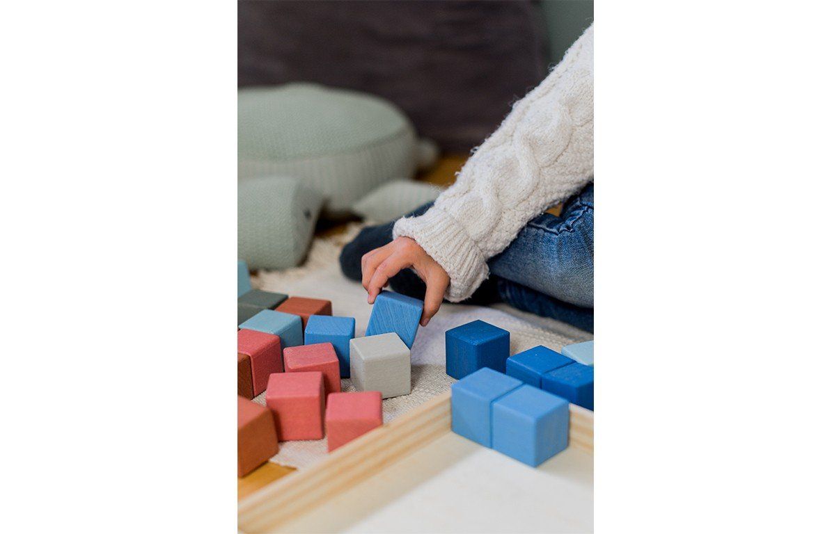 Kind greift nach einem blauen Holzbaustein. Mehrere Bausteine in verschiedenen Farben liegen auf dem Boden.