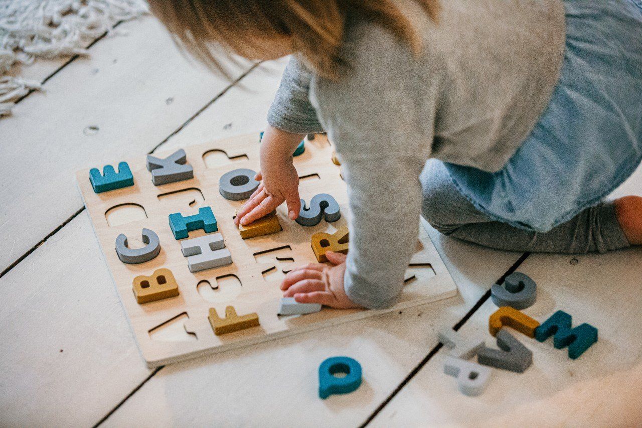 Kind spielt mit einem Holzpuzzle. Das Puzzle hat Buchstaben in verschiedenen Farben. Das Kind sitzt auf dem Boden.
