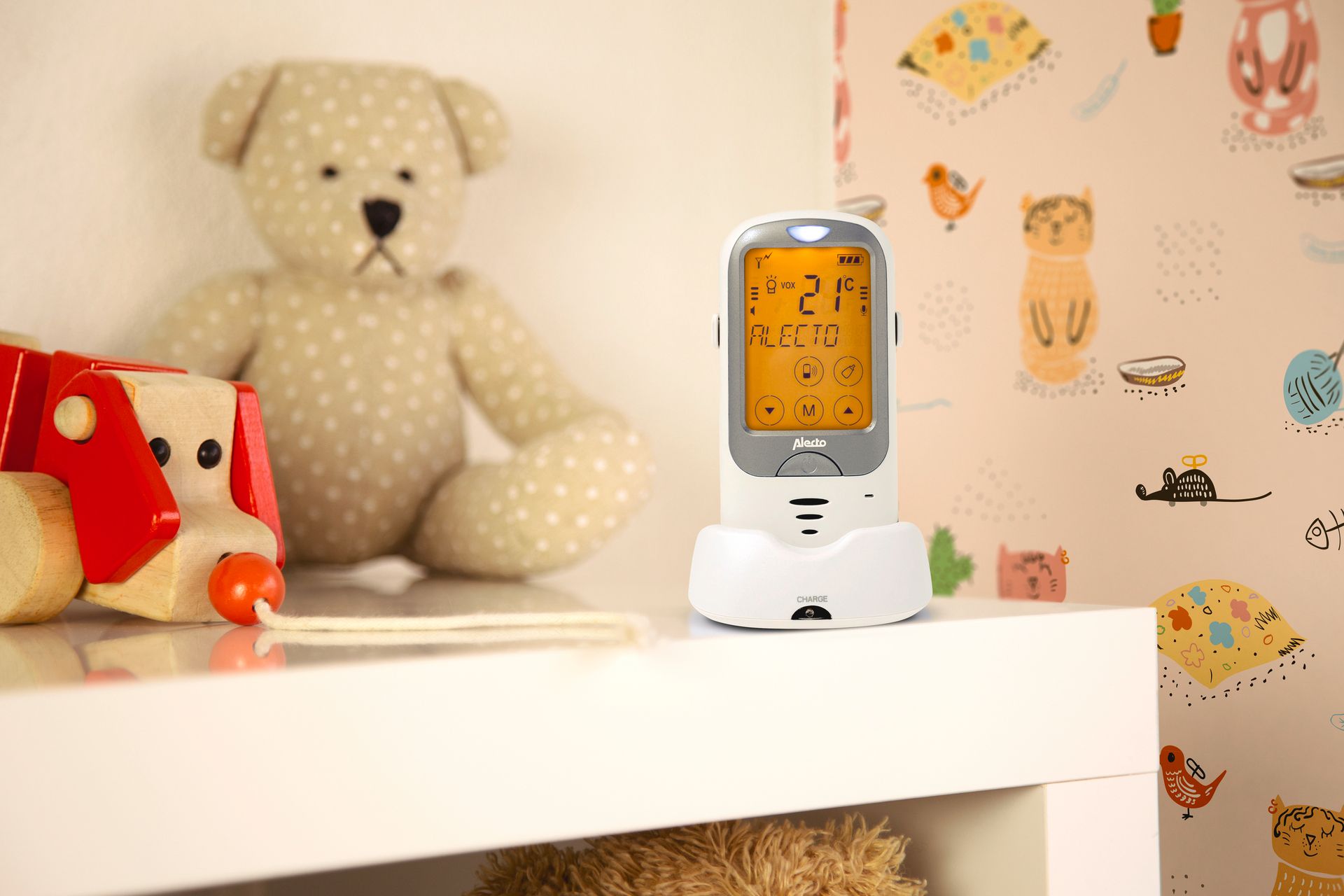 Babyphone auf einem weißen Regal. Das Display zeigt Informationen wie Temperatur und den Markennamen ALECTO an. Im Hintergrund Spielzeug und Tapete.