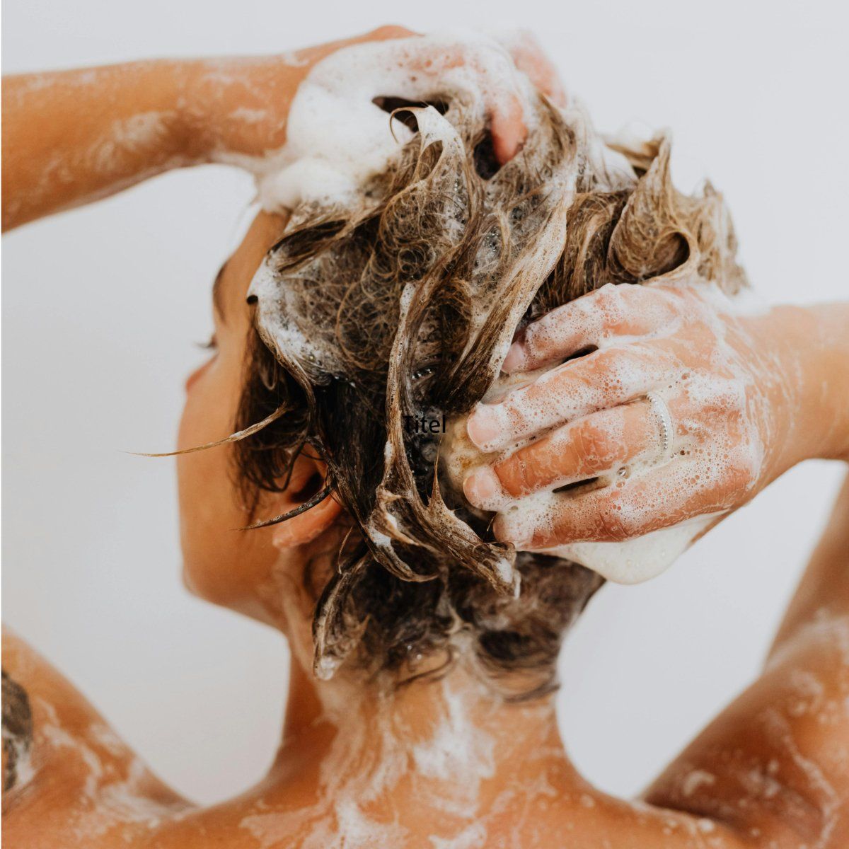 Person wäscht Haare mit Schaum. Hände massieren Kopfhaut. Rückansicht, heller Hintergrund.