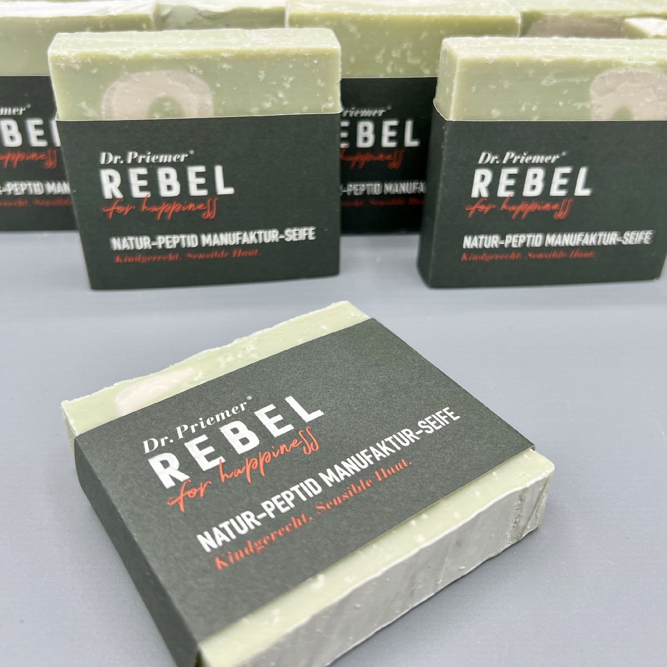 Mehrere Seifen mit dunkler Banderole. Aufschrift: Dr. Priemer, REBEL for happiness, Natur-Peptid Manufaktur-Seife.
