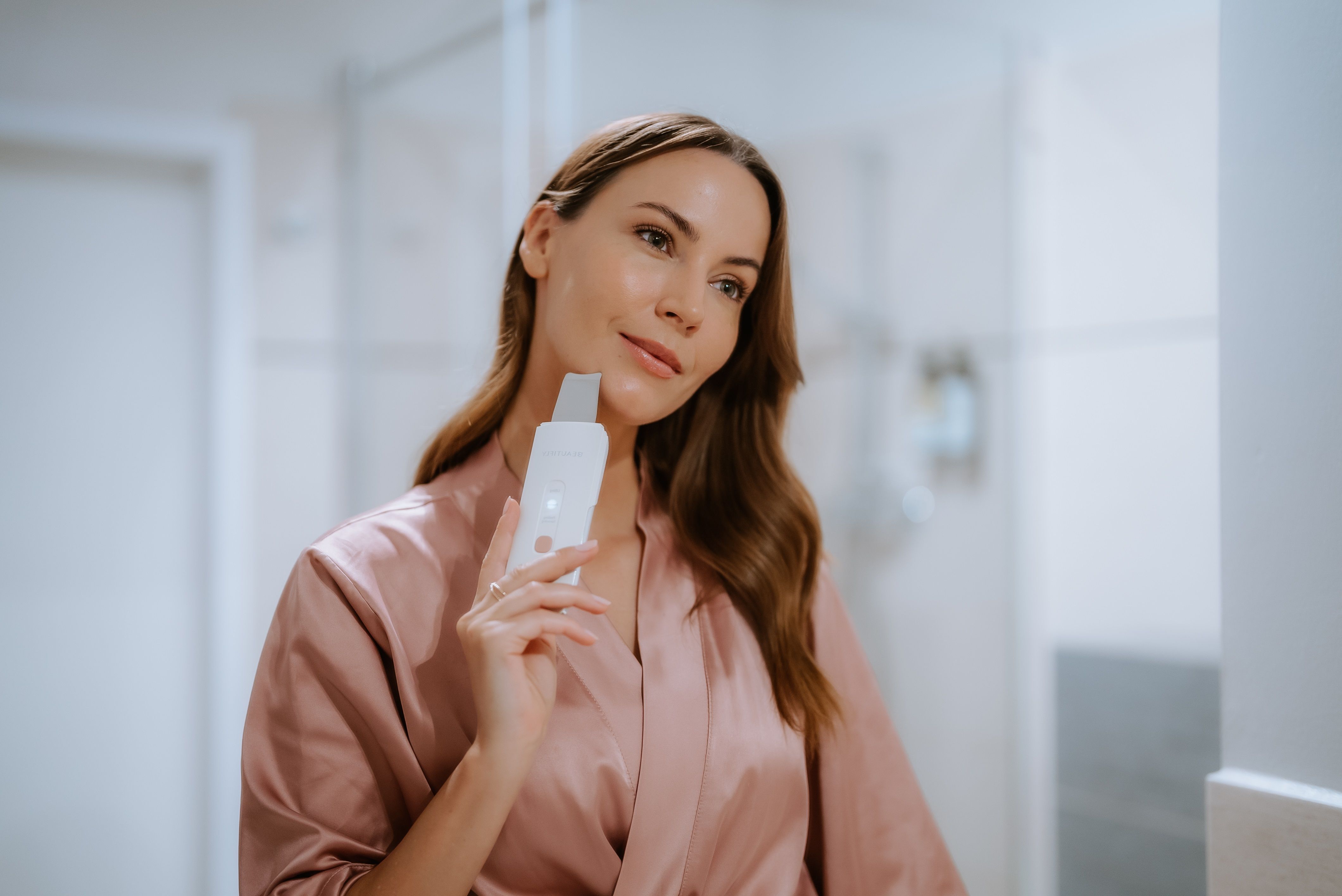 Frau hält ein Gerät vor dem Spiegel. Gerät beige mit Metallklinge. Frau trägt einen rosa Bademantel.