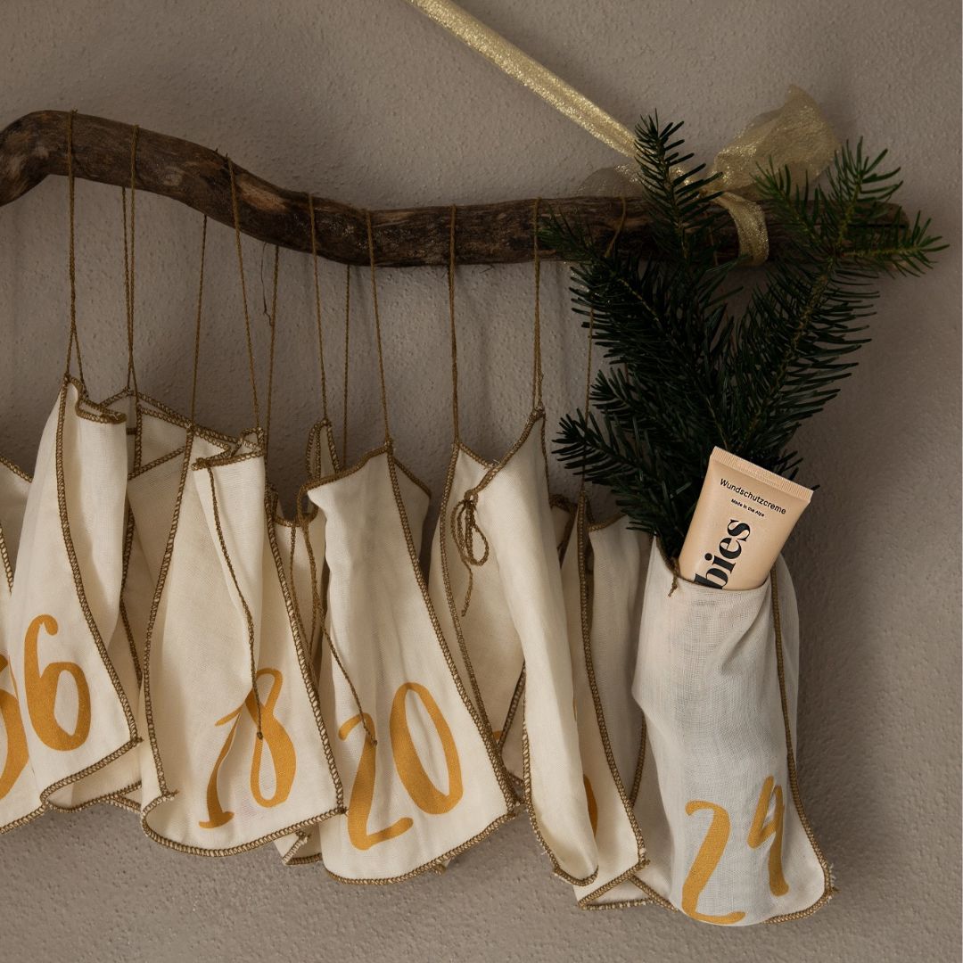 Adventskalender mit Stoffbeuteln, gefüllt mit Produkten. Eine Tube Creme ist sichtbar. Auf einem Ast mit Tannenzweig.