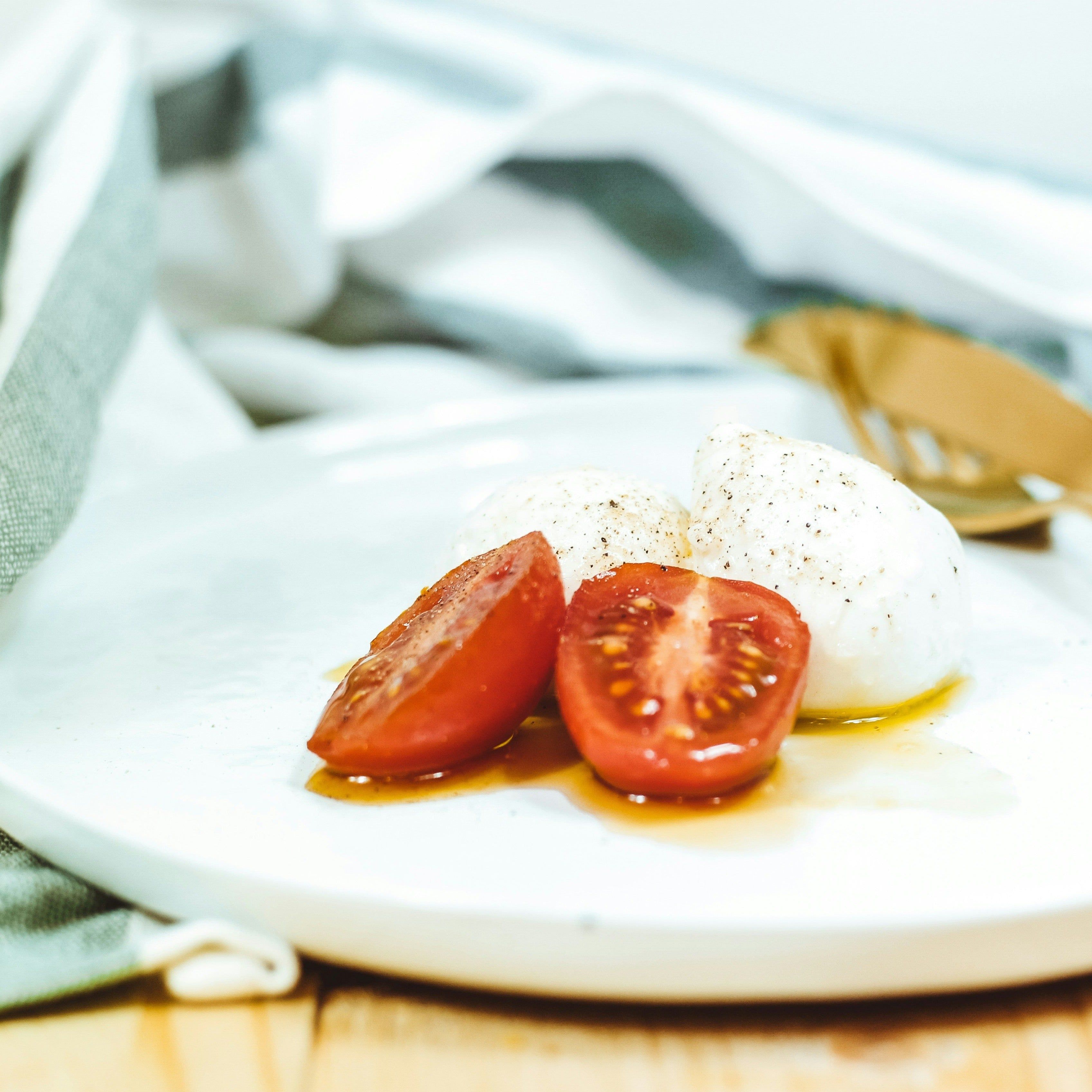 Teller mit Tomatenhälften und Mozzarella-Kugeln. Gewürze und Öl. Hintergrund: Stoffserviette und Holzoberfläche.
