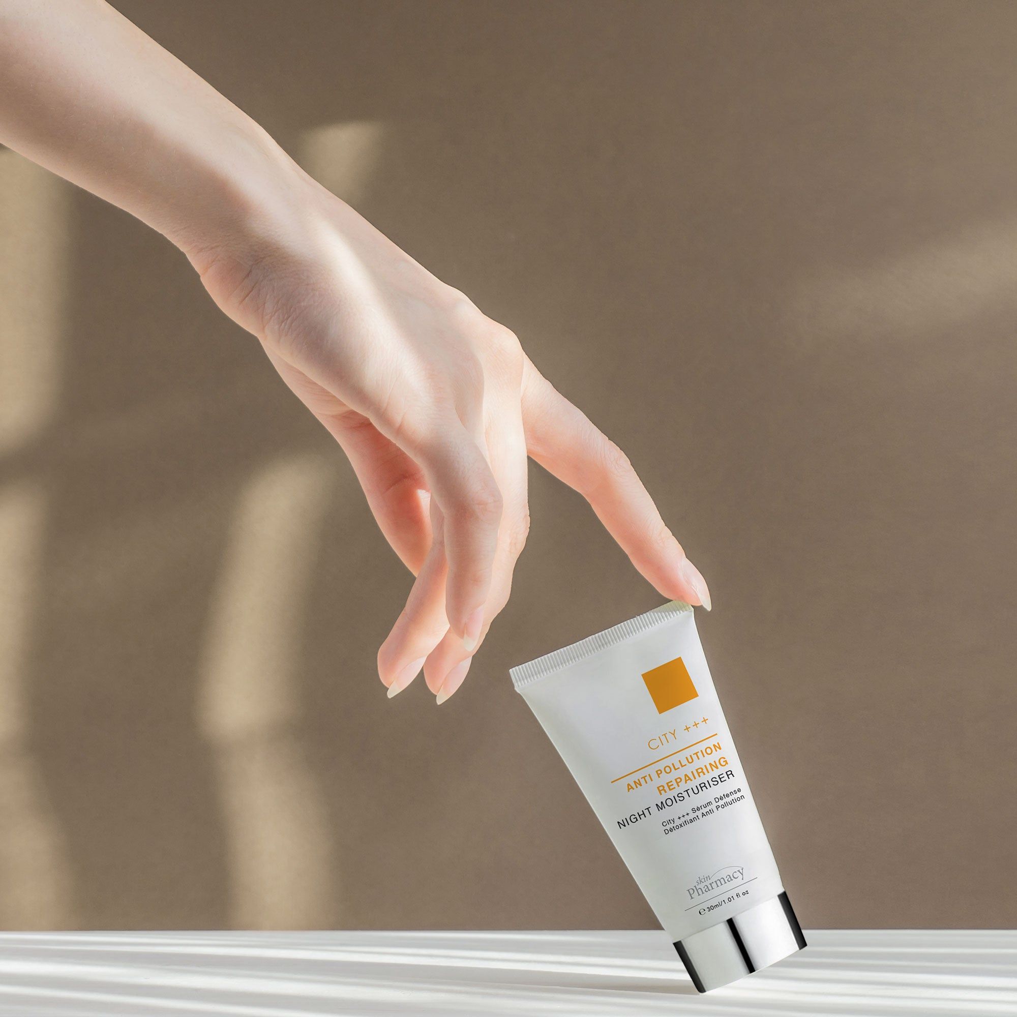 Weiße Tube, silberner Deckel. Aufschrift: ANTI POLLUTION REPAIRING NIGHT MOISTURISER. Marke: skinPharmacy. Hand berührt die Tube.