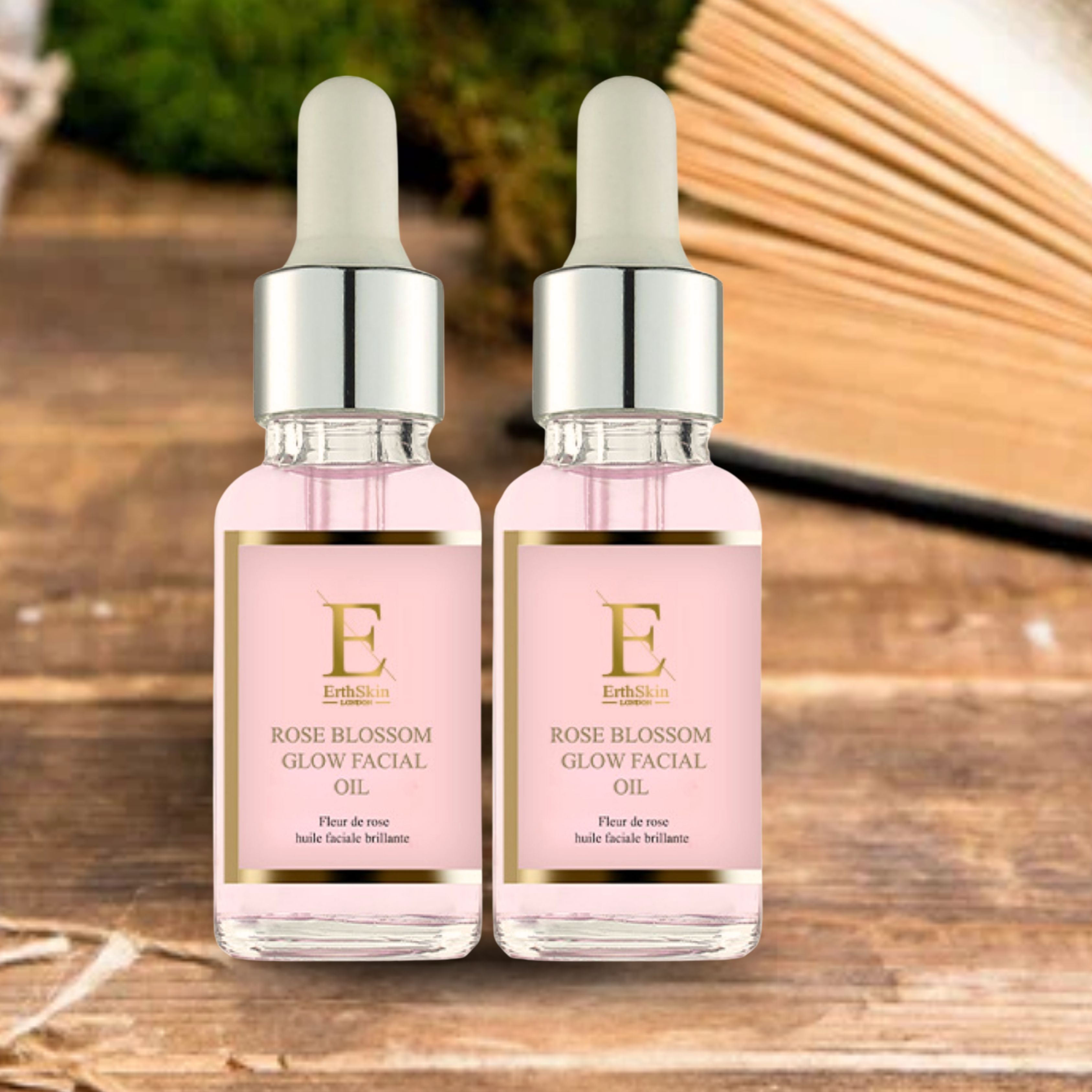 Deux flacons d'huile pour le visage Rose Blossom Glow. Liquide rose dans des flacons en verre avec pipettes argentées. Étiquette avec logo et texte, devant un livre.