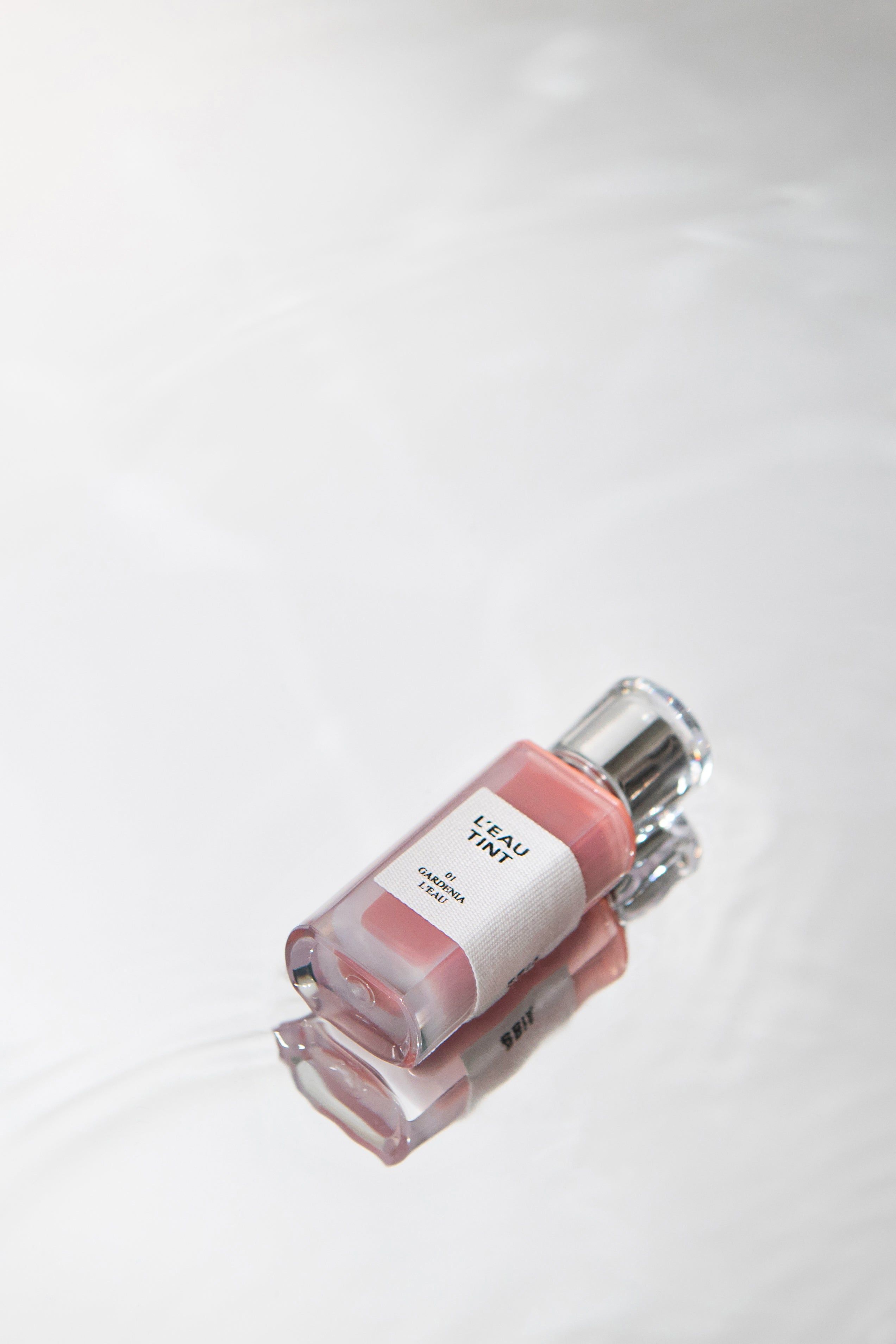 Flasche Lippen-Tönungsmittel auf spiegelnder Oberfläche. Aufkleber mit "L'EAU TINT" und "GARDENIA L'EAU".