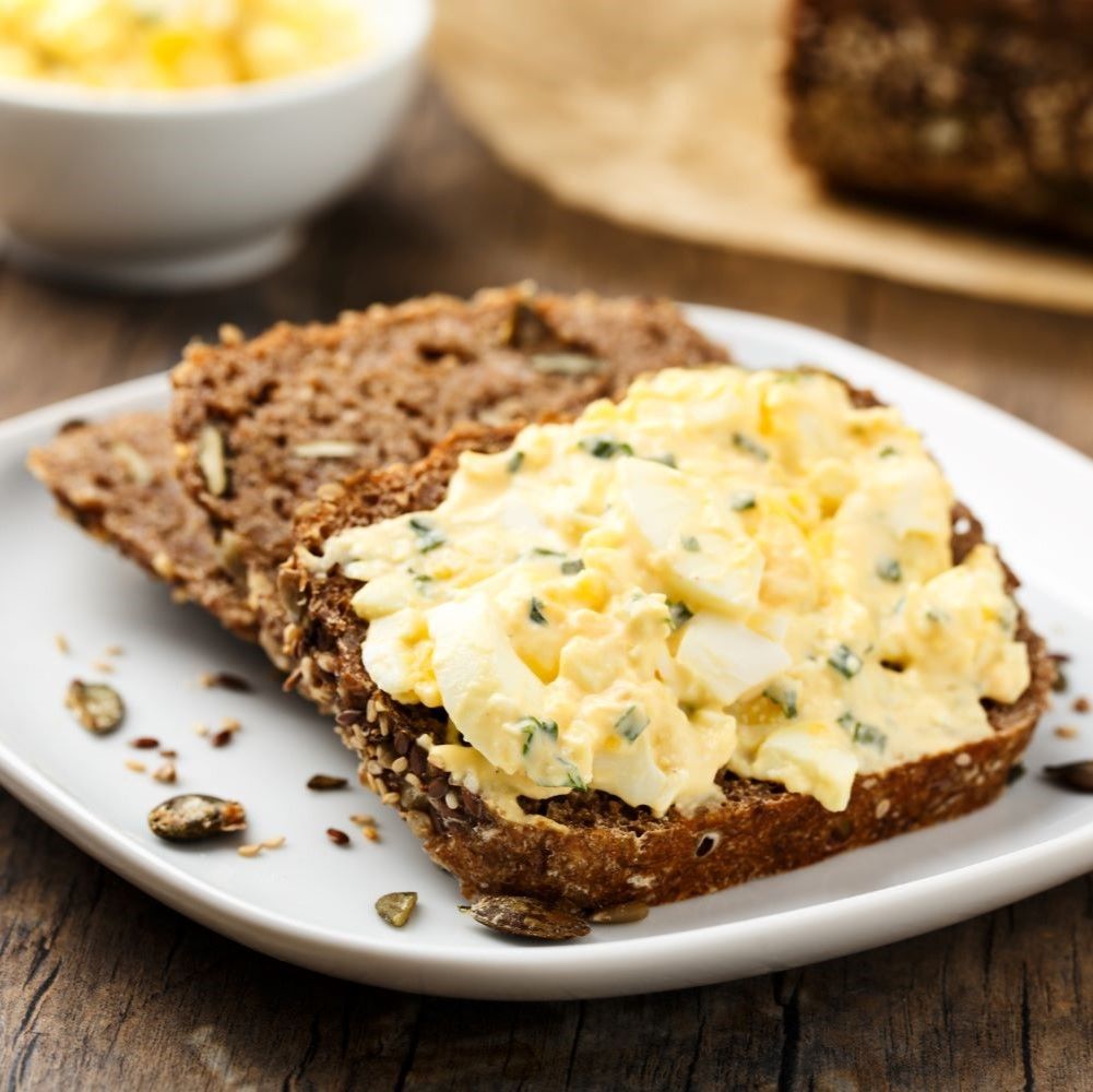 Scheiben Brot mit Eiersalat auf einem Teller. Daneben Brot und Samen. Auf einem Holztisch.