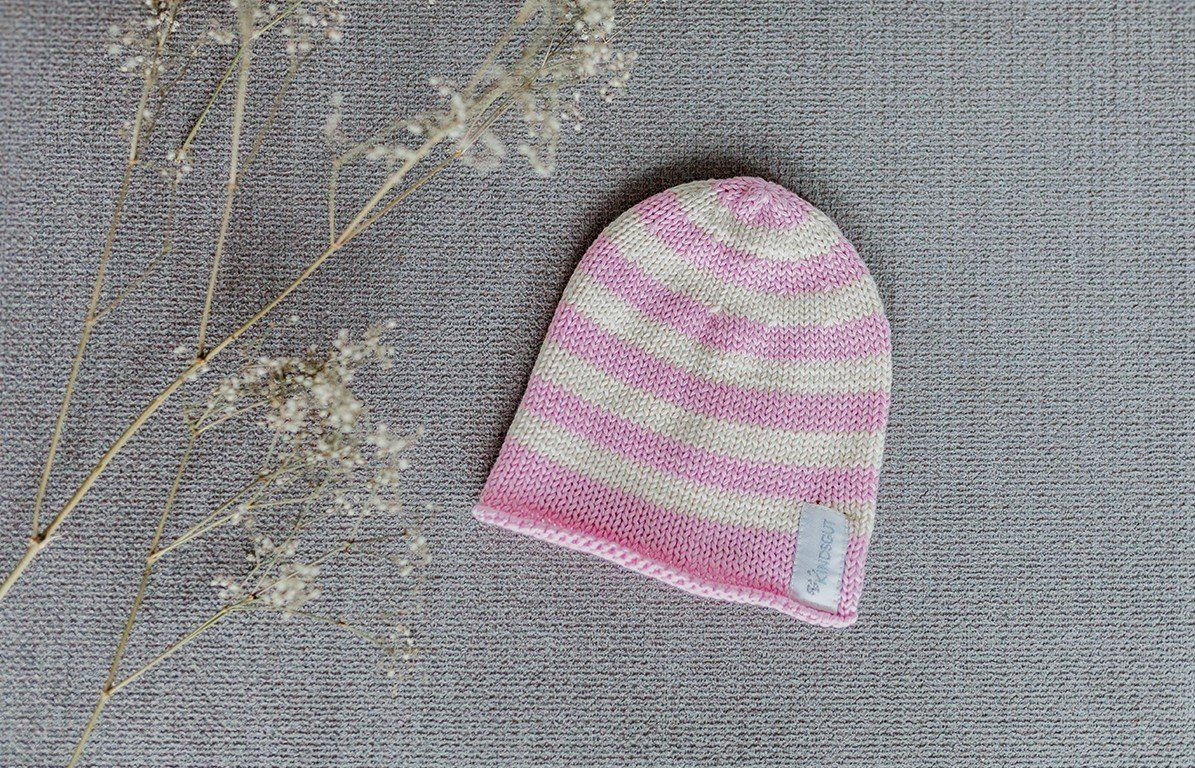 Rosa-weiß gestreifte Kindermütze. Label mit Kindsgut-Logo. Auf grauem Untergrund.
