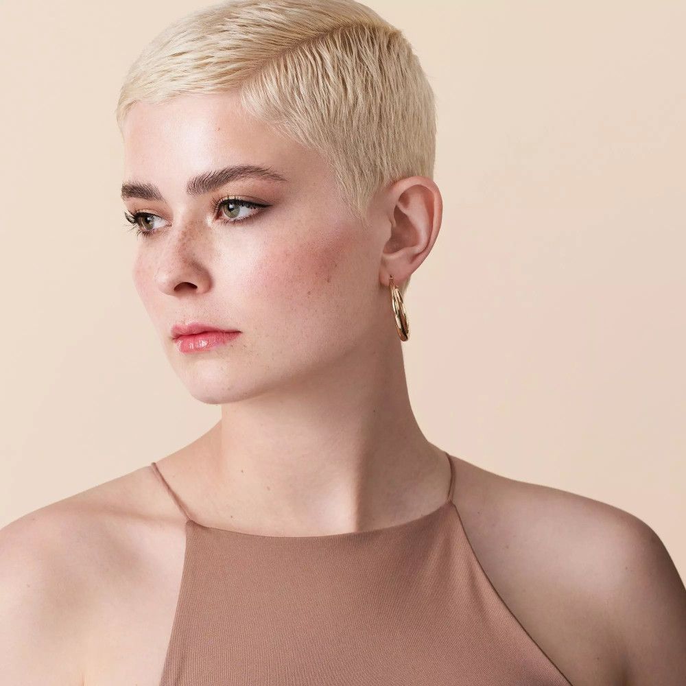 Porträt einer Frau mit kurzen blonden Haaren. Sie trägt ein braunes Oberteil und Ohrringe.