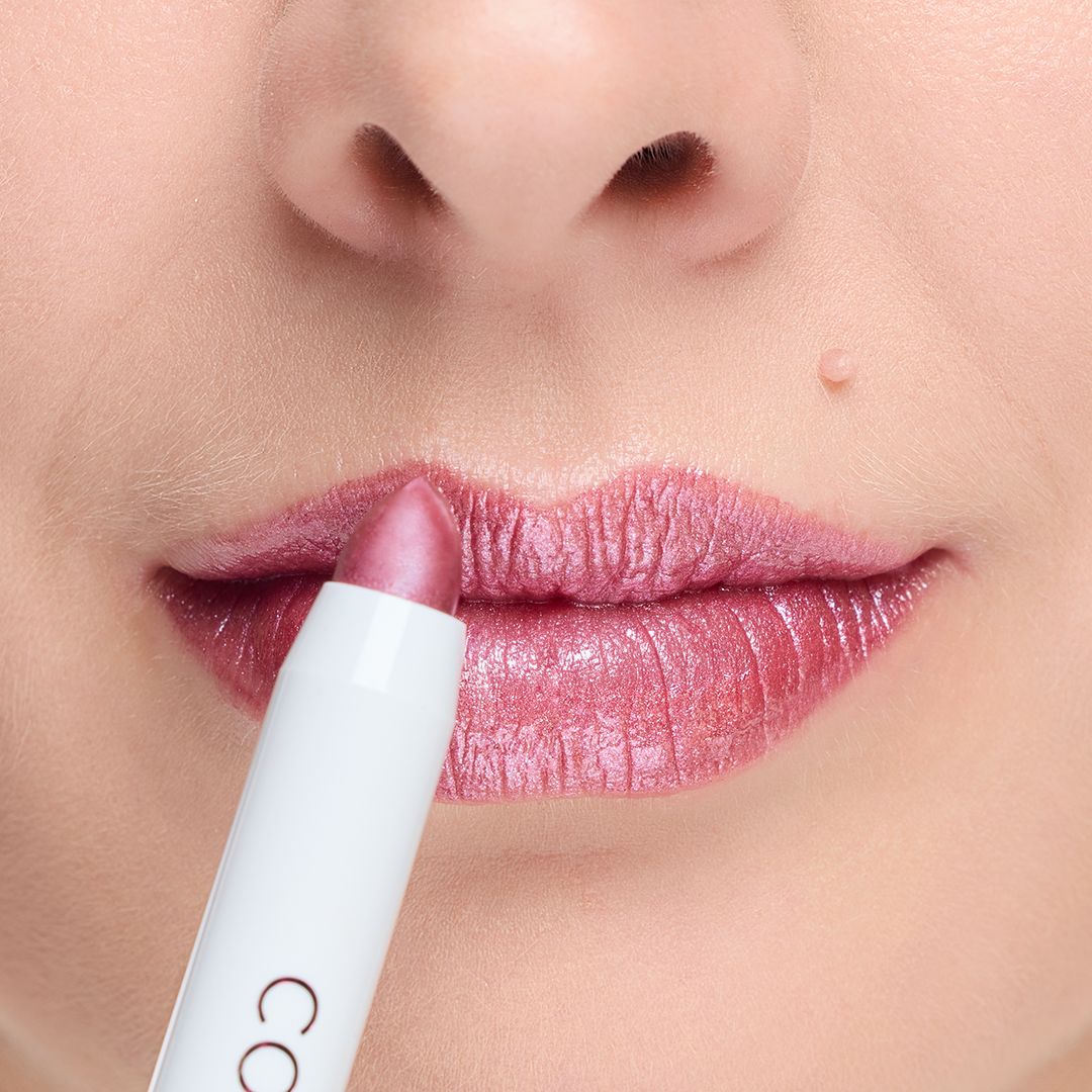 Lippenstift wird auf Lippen aufgetragen. Der Lippenstift ist rosa und glänzend. Der Lippenstift hat eine weiße Hülle mit dem Markennamen.
