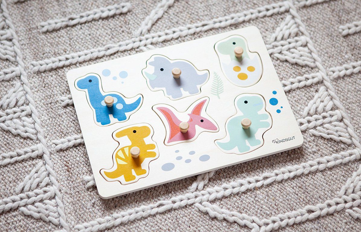 Holzpuzzle mit sechs bunten Dinosauriern auf einem grauen Strickmuster. Knöpfe an den Teilen. Kindsgut-Logo.