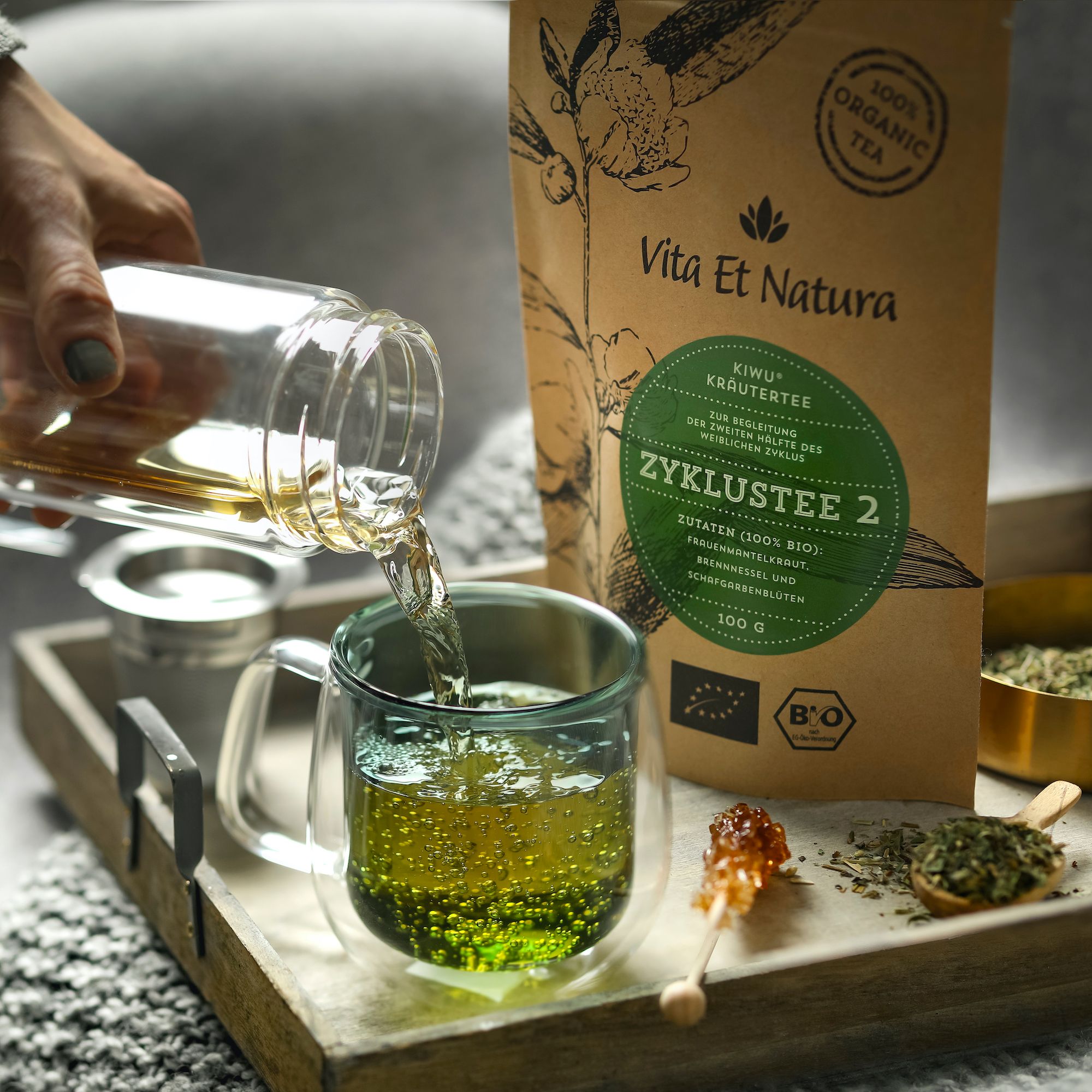 Tee wird in eine Tasse gegossen. Daneben Beutel mit Zyklustee 2. Aufschrift: Vita Et Natura.
